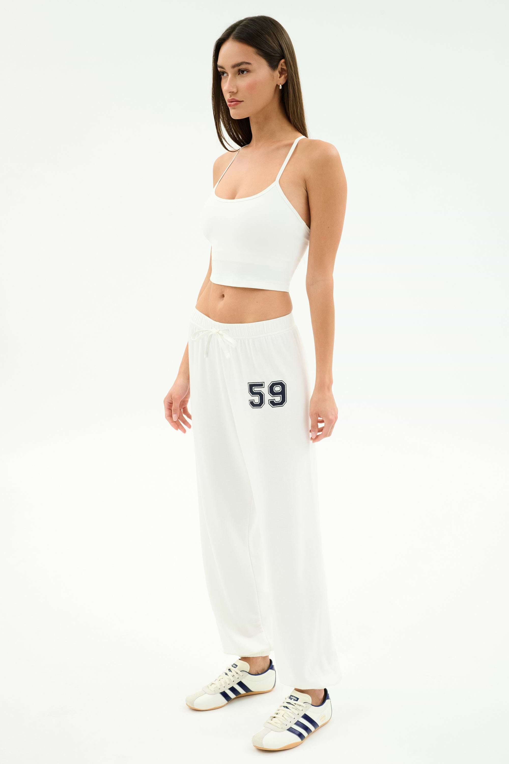A woman in a white studio wears a white crop top and the SPLITS59 Andie Oversized Fleece Varsity 59 Sweatpant (White/59), ideal for cool weather workouts, finished with white sneakers with navy stripes.