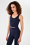 A woman with curly hair wears the SPLITS59 Sprint Rigor Bralette in Indigo, styled for multi-sport performance, standing against a plain white background.
