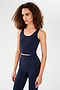 A woman with curly hair wears the SPLITS59 Sprint Rigor Bralette in Indigo, styled for multi-sport performance, standing against a plain white background.