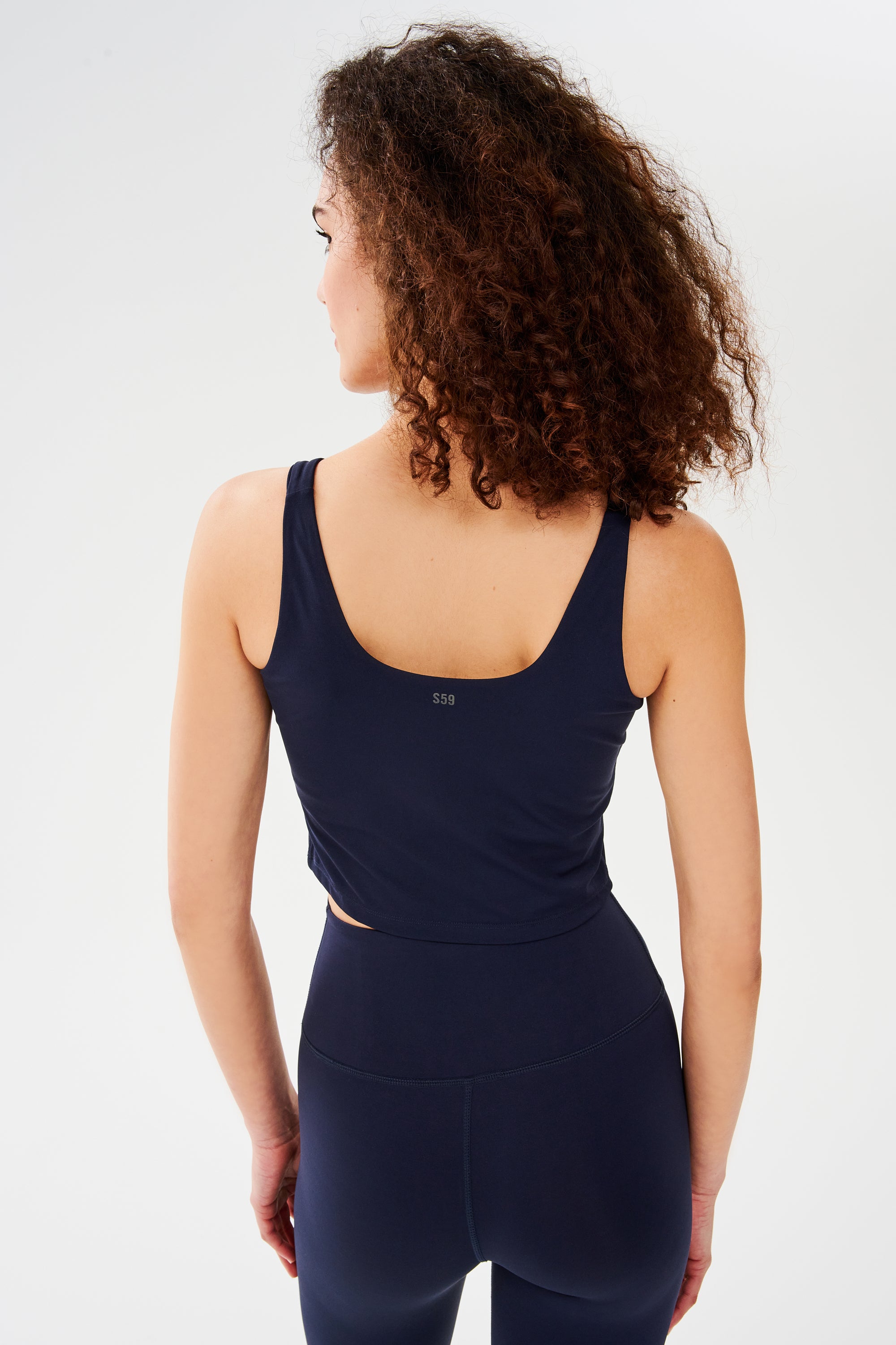 A woman with curly hair is seen from the back against a white background, wearing the SPLITS59 Sprint Rigor Bralette in Indigo, designed for multi-sport performance.
