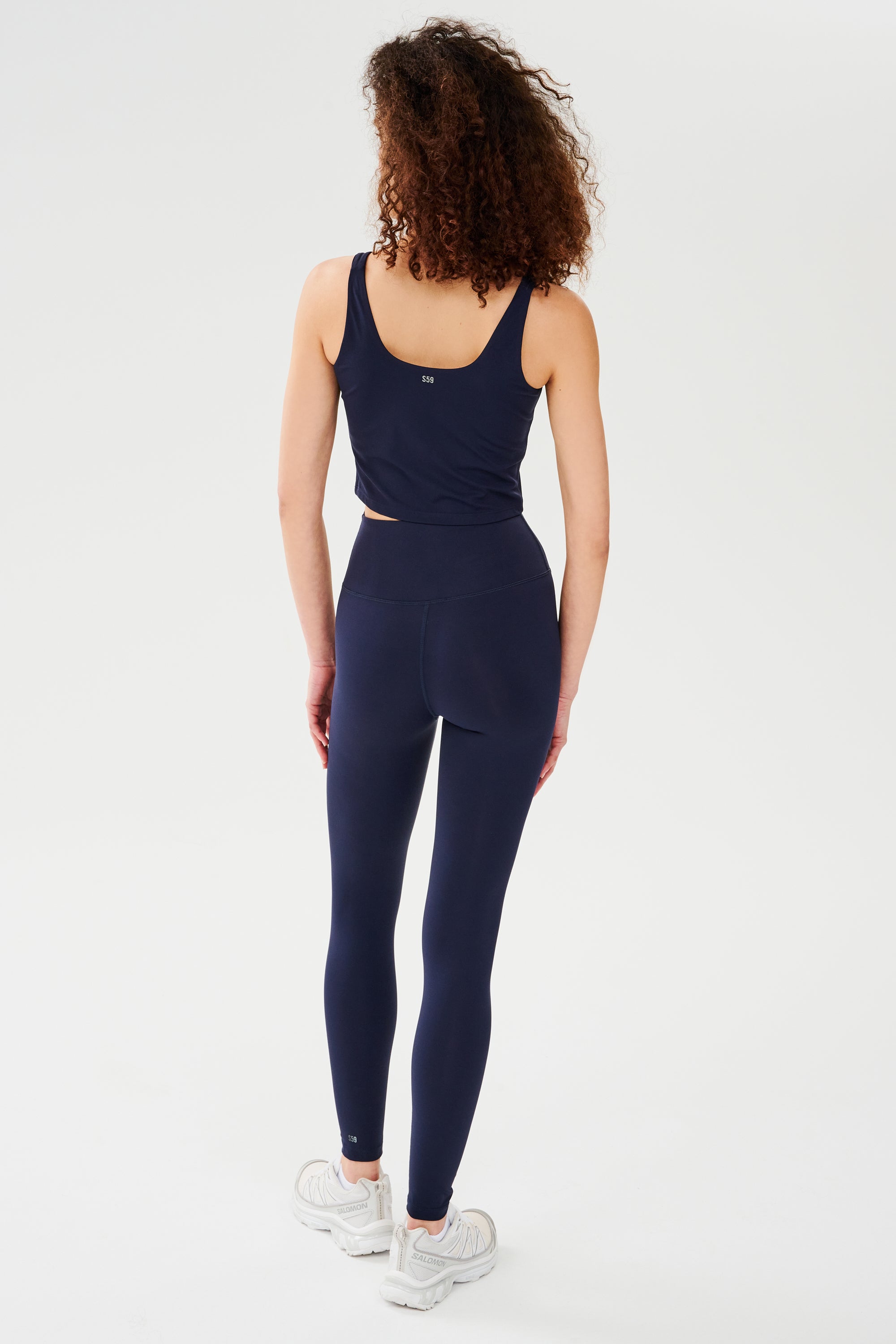 A person with curly hair wears the SPLITS59 Sprint Rigor Bralette in Indigo along with leggings and white sneakers, standing facing away on a plain white background.
