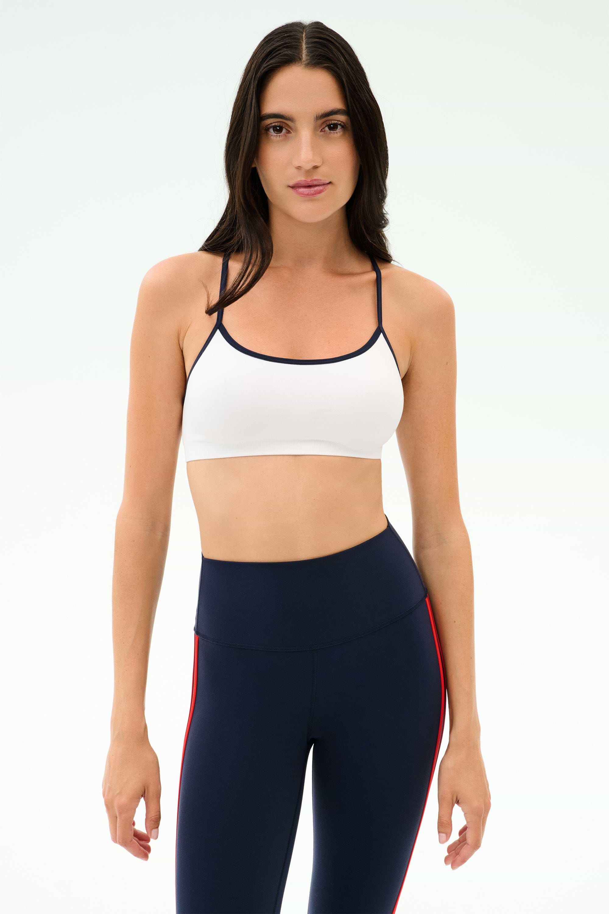 A woman stands against a plain background wearing the SPLITS59 Loren Seamless Bra in White/Indigo, perfect for gym workouts, paired with navy blue athletic leggings featuring red stripes.