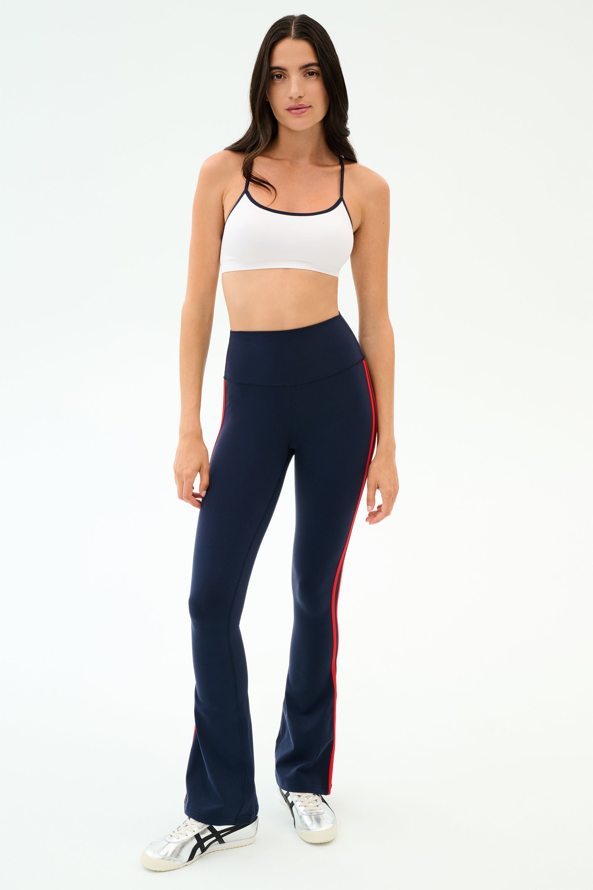 A woman faces forward against a plain background, wearing SPLITS59's Raquel High Waist Flared Legging in Indigo/Lotus Red and a white sports bra with white sneakers.