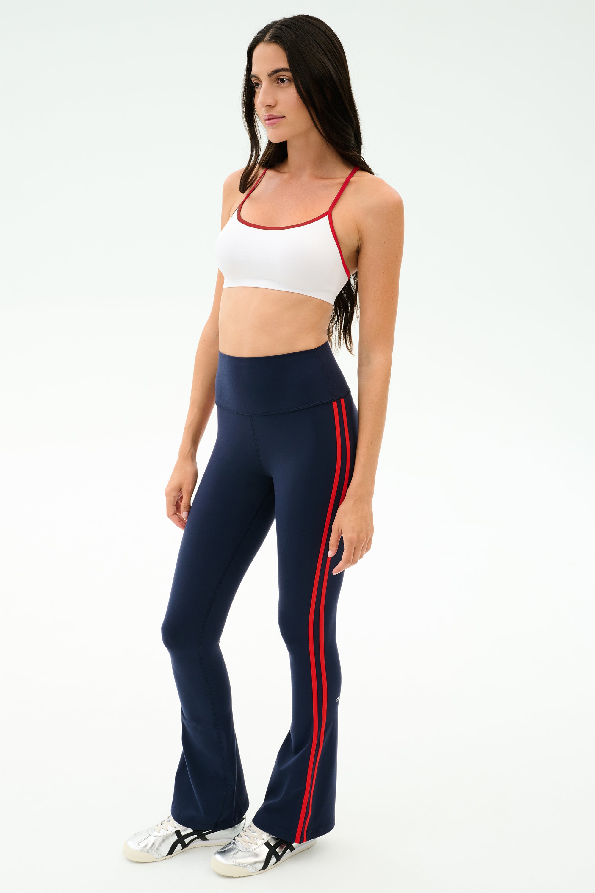 A woman poses in a studio wearing SPLITS59's Raquel High Waist Flared Legging in Indigo/Lotus Red and a white sports bra, paired with white sneakers against a plain background.