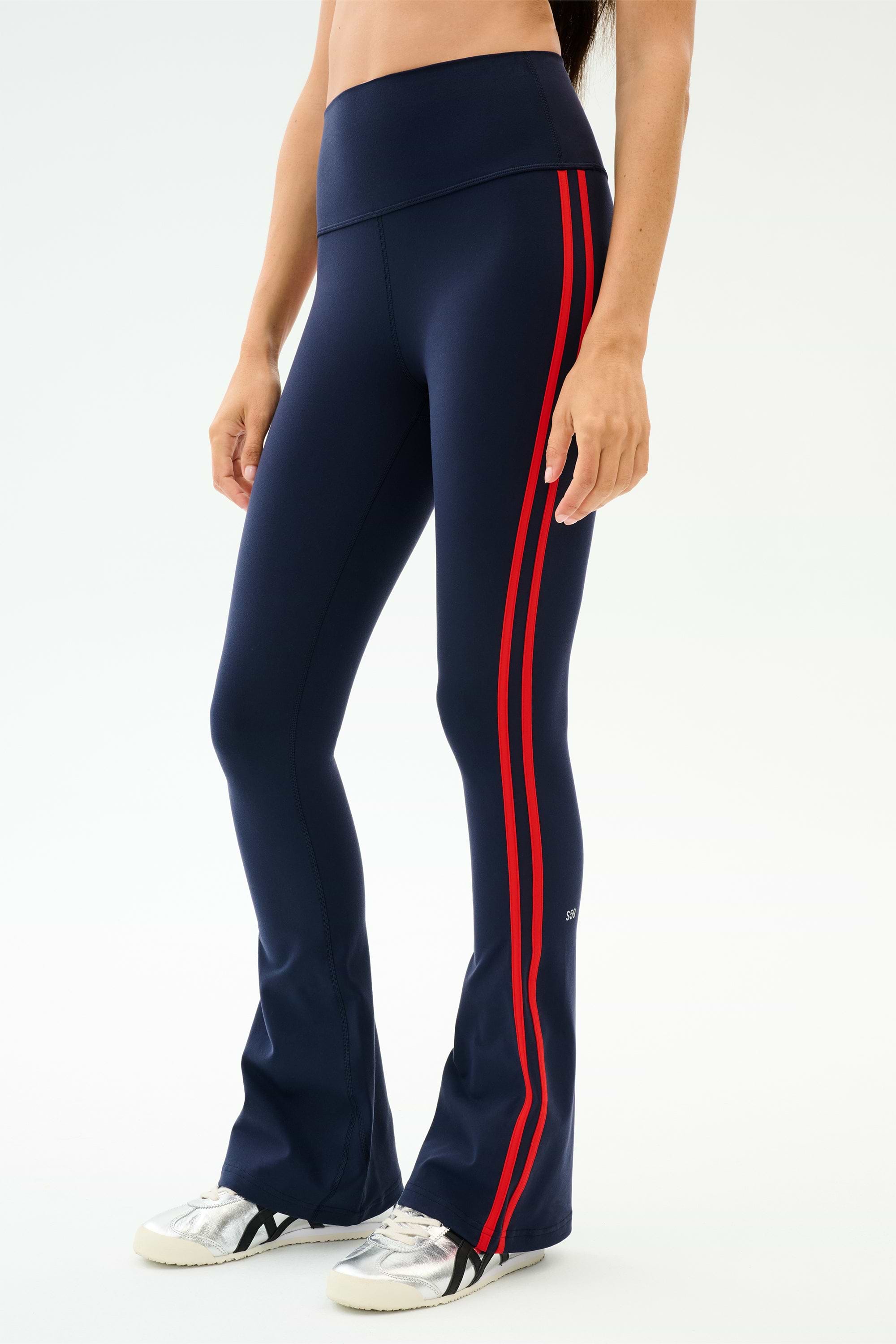 A person wears the SPLITS59 Raquel High Waist Flared Legging in Indigo/Lotus Red with white sneakers, standing against a plain white background. These stylish leggings offer comfortable 4-way stretch for easy movement.