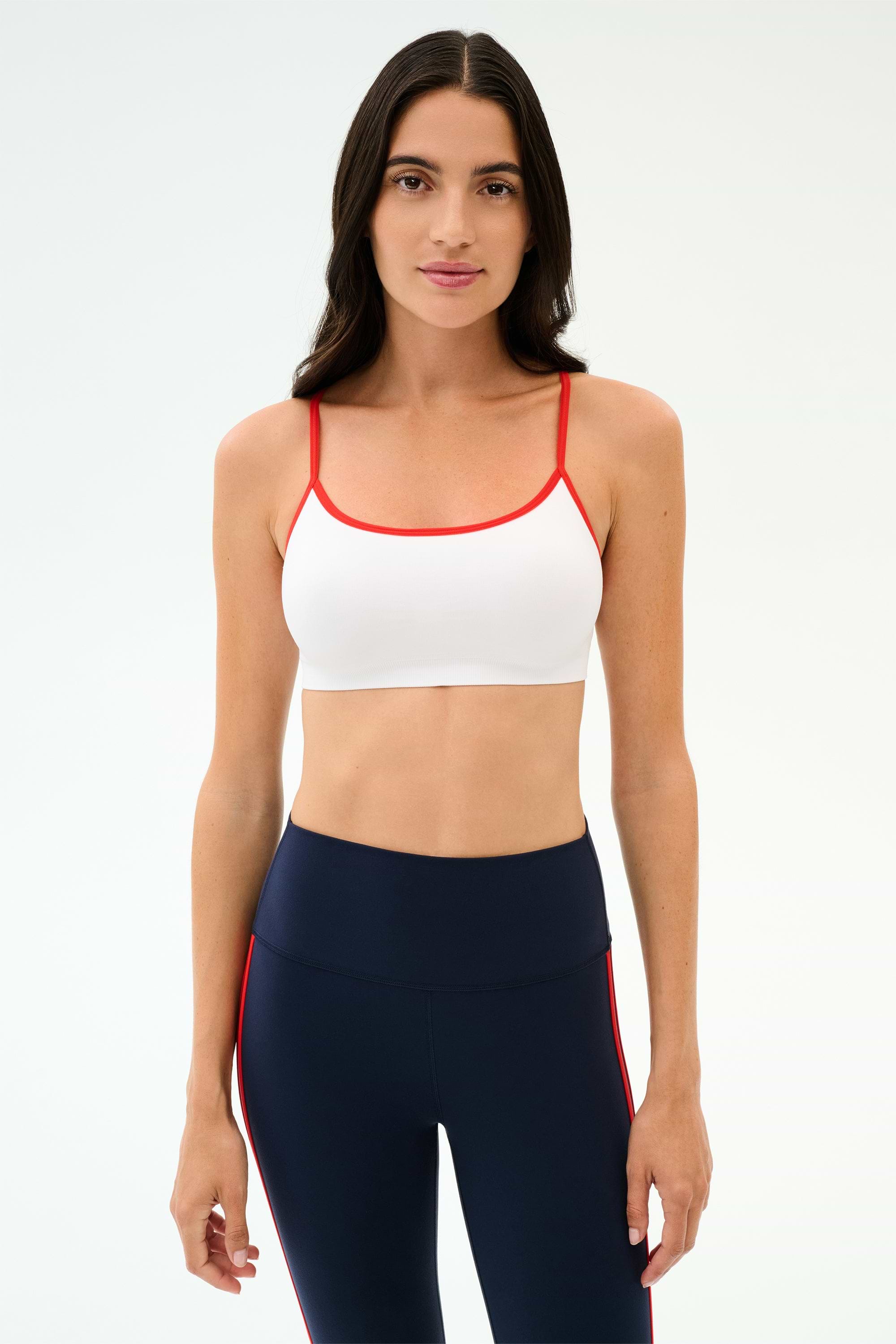 A woman faces forward in a SPLITS59 Loren Seamless Bra - White/Lotus Red, perfect for workouts, paired with high-waisted navy leggings featuring red side stripes, set against a plain light background.