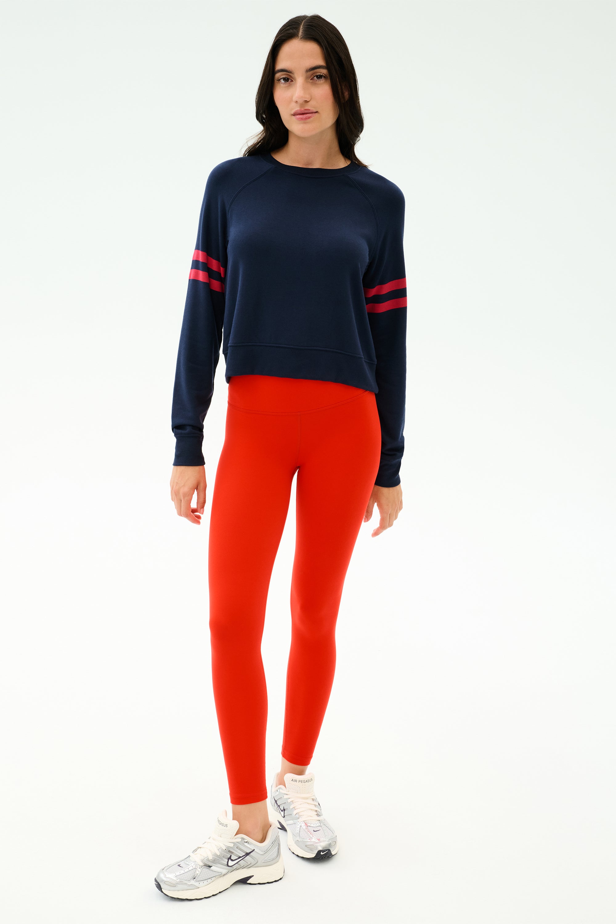 A woman stands against a plain background, wearing the SPLITS59 Brooks Stripe Fleece Sweatshirt in Indigo/Lotus Red, bright red leggings, and white sneakers.