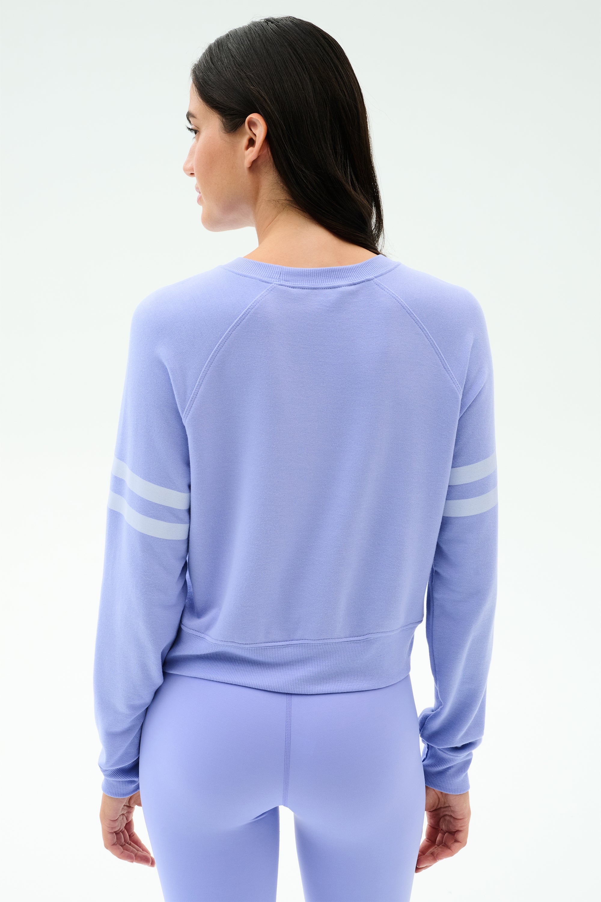 A woman with long dark hair wears the SPLITS59 Brooks Stripe Fleece Sweatshirt in Purple Haze/White, featuring two white sleeve stripes, made in Los Angeles, standing with her back to the camera.