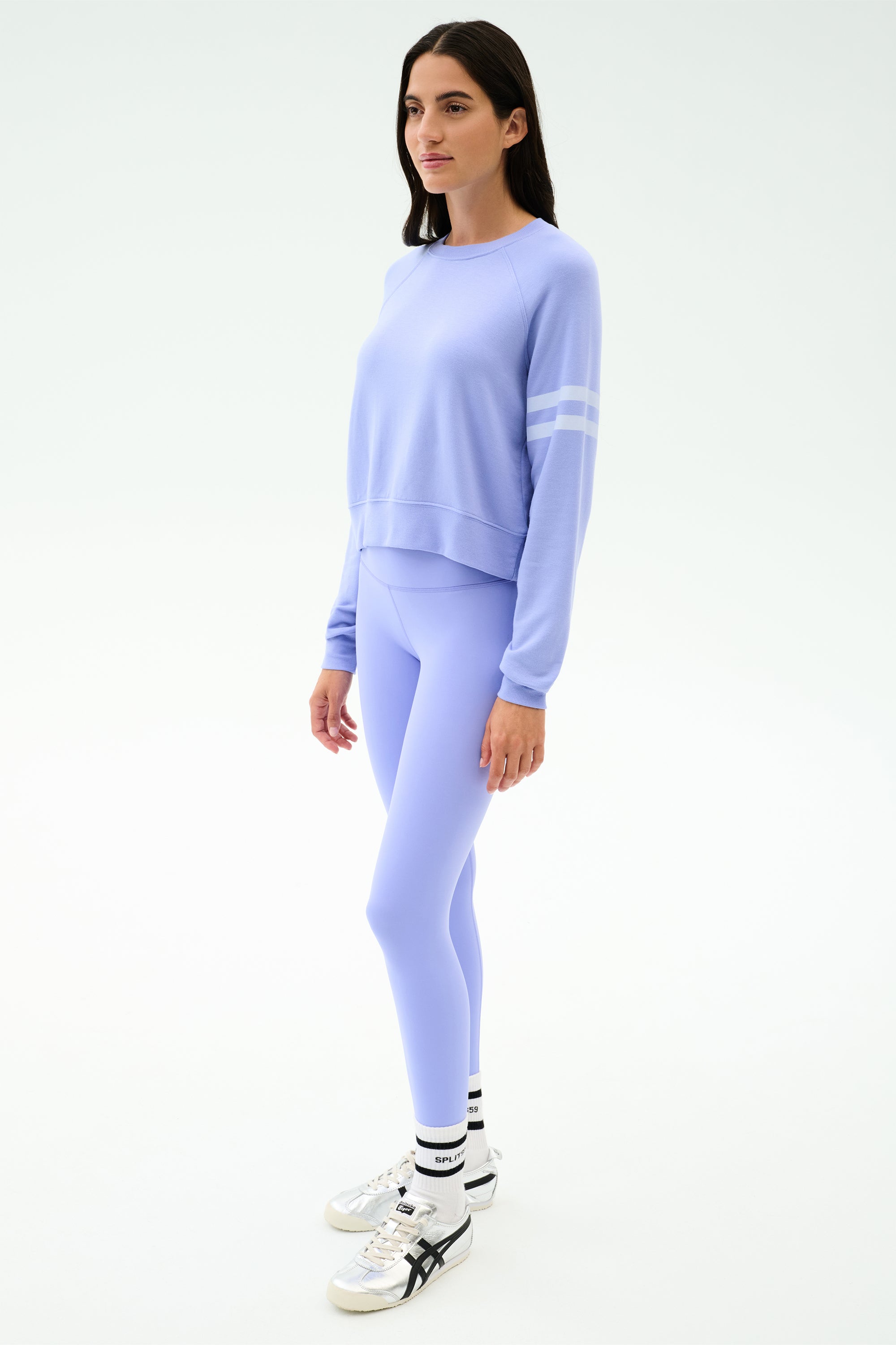 A woman faces slightly left, wearing the SPLITS59 Brooks Stripe Fleece Sweatshirt in Purple Haze/White with matching leggings, white socks, and sneakers against a plain light background.