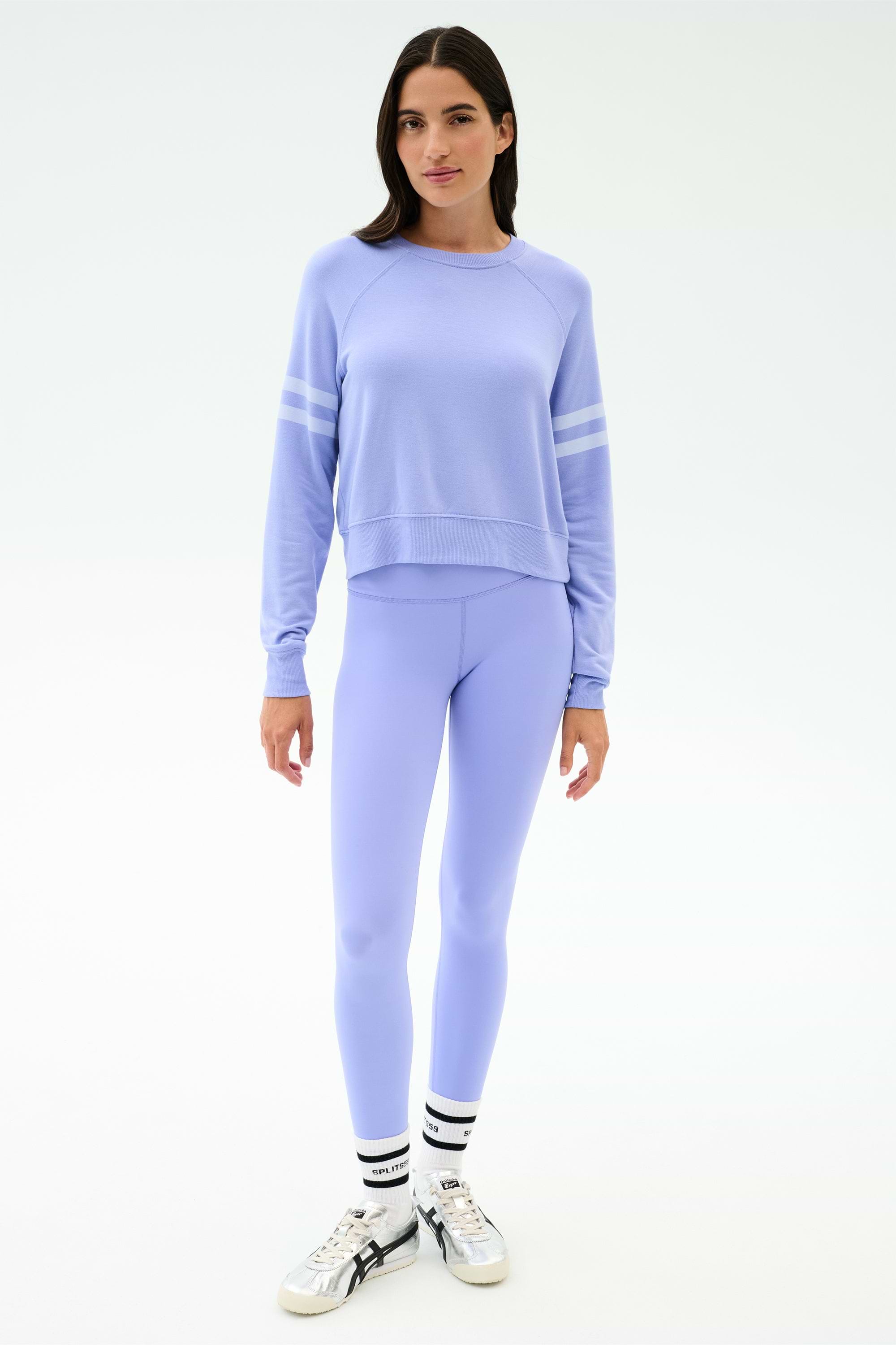 A woman stands facing forward, wearing the SPLITS59 Brooks Stripe Fleece Sweatshirt in Purple Haze/White with matching leggings, white socks with black stripes, and white sneakers, set against a plain white background.