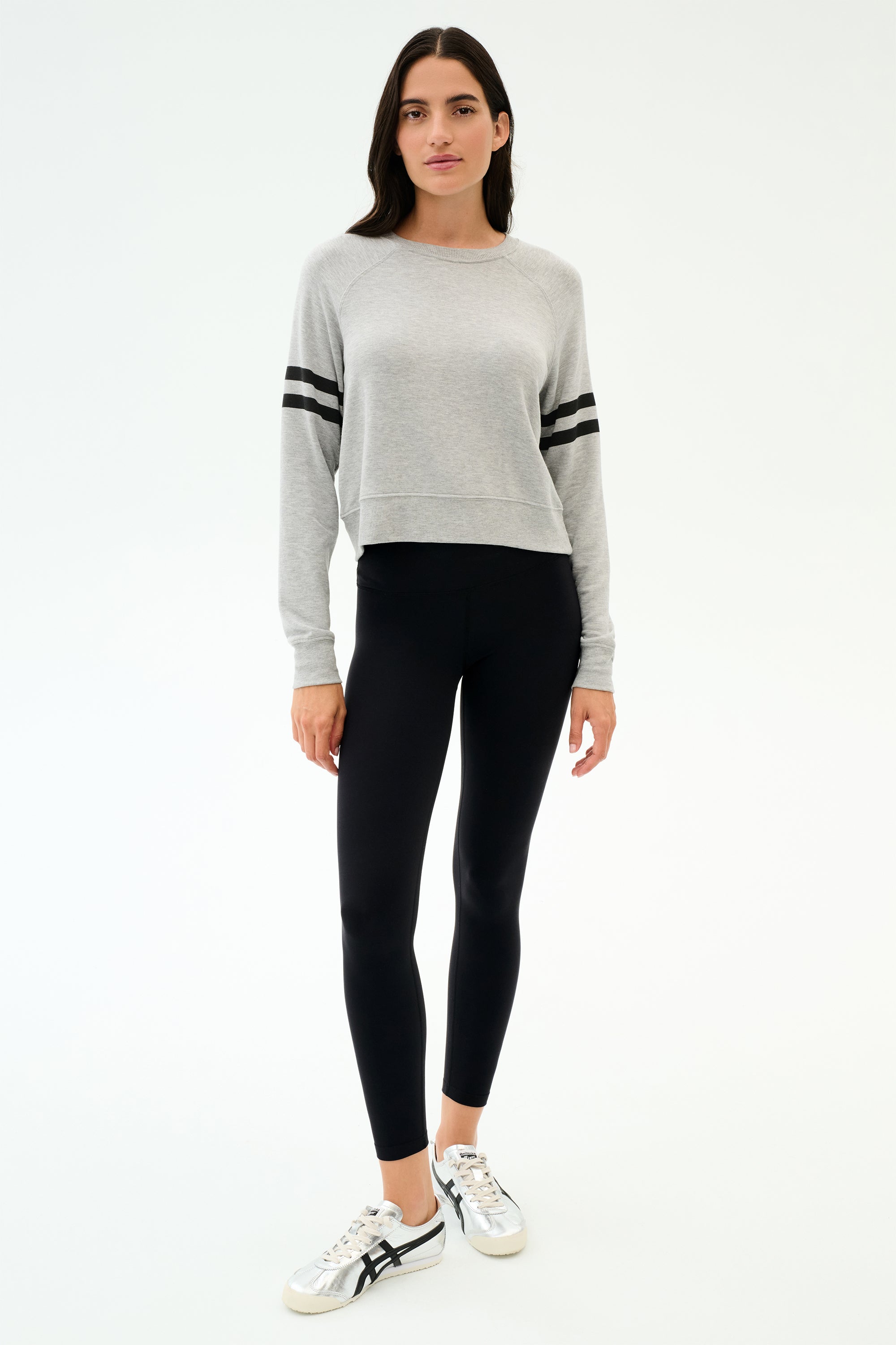 A person stands facing forward wearing the SPLITS59 Brooks Stripe Fleece Sweatshirt in Heather Grey/Black, paired with black leggings and white sneakers, set against a plain white background.
