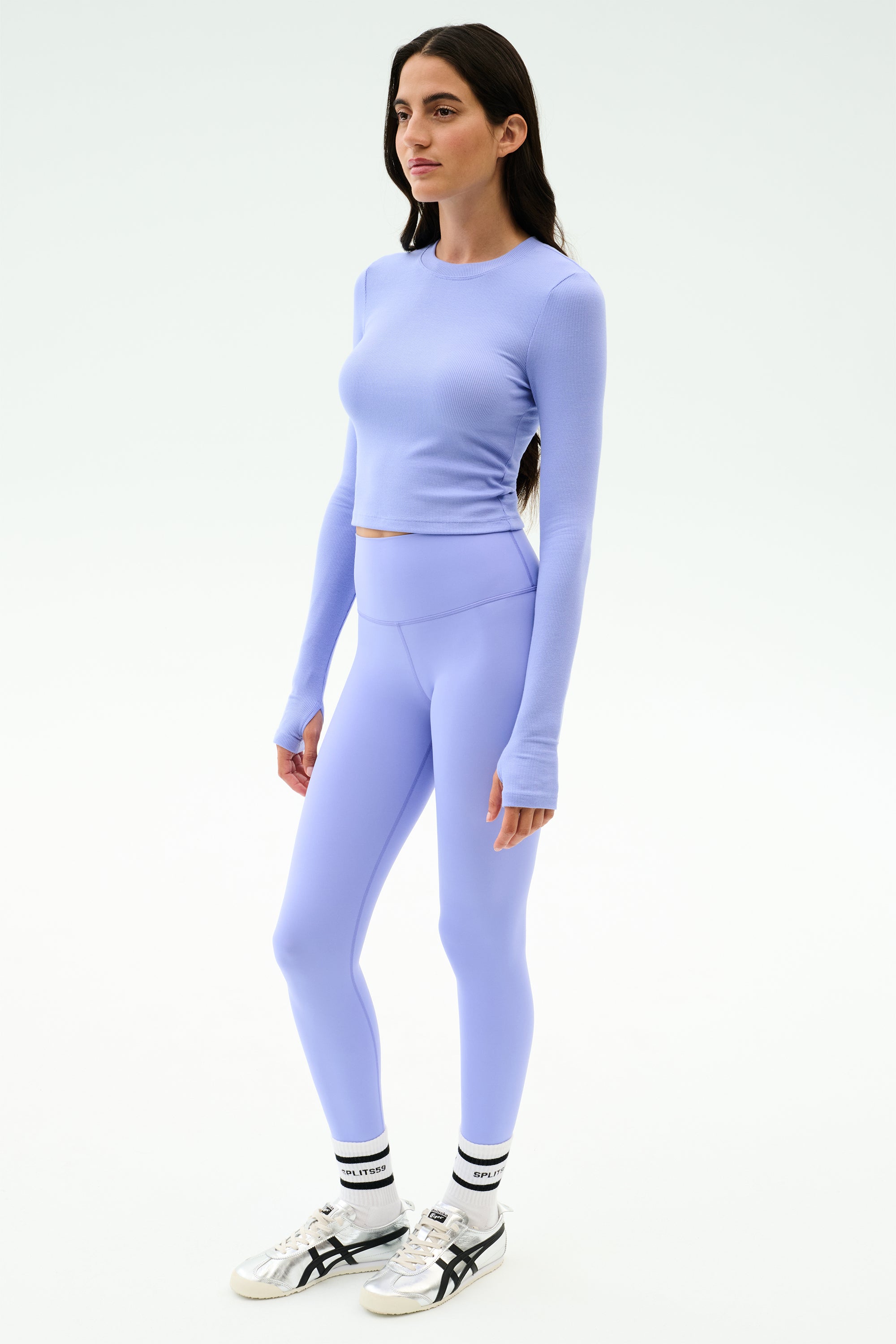 A woman stands against a white background wearing the SPLITS59 Louise Rib Long Sleeve Crop in Purple Haze, paired with matching leggings, white socks, and white sneakers.