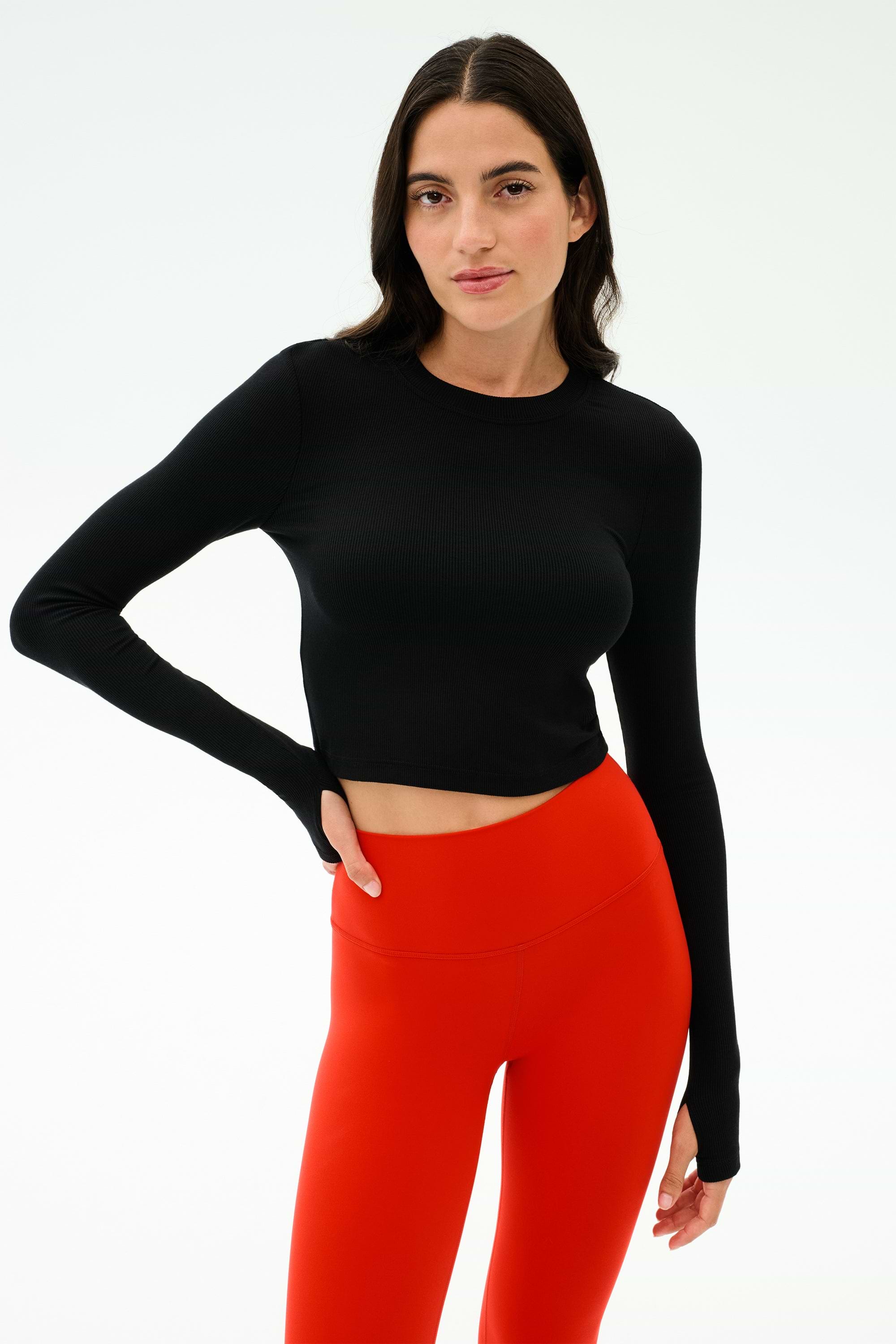 A woman wears the SPLITS59 Louise Rib Long Sleeve Crop in black with high-waisted red leggings, standing against a plain light background.