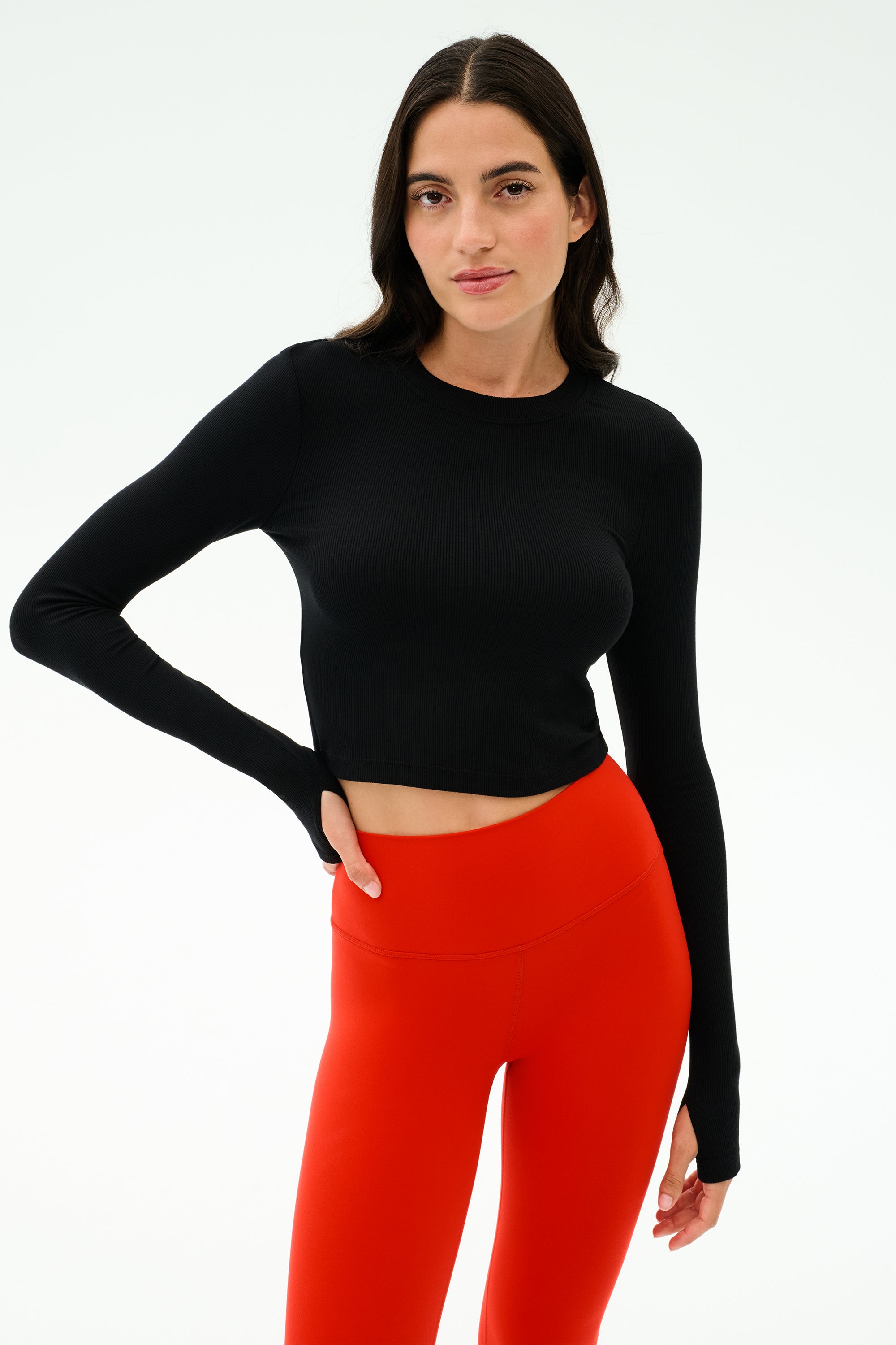 A woman wears the SPLITS59 Louise Rib Long Sleeve Crop in black with high-waisted red leggings, standing against a plain light background.