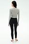 A woman faces away, wearing the SPLITS59 Louise Rib Long Sleeve Crop in Heather Grey, paired with black high-waisted leggings and white athletic shoes, set against a plain light background.
