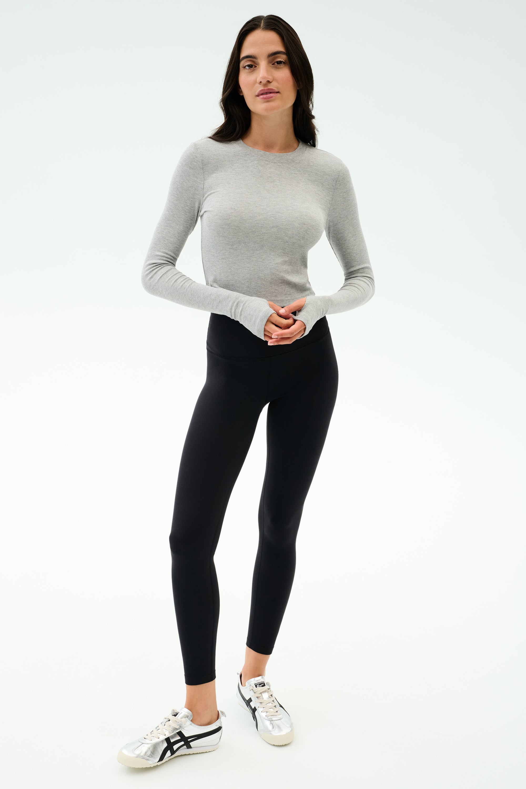 A woman stands against a white background wearing the SPLITS59 Louise Rib Long Sleeve Crop in Heather Grey, black leggings, and white sneakers with black accents.
