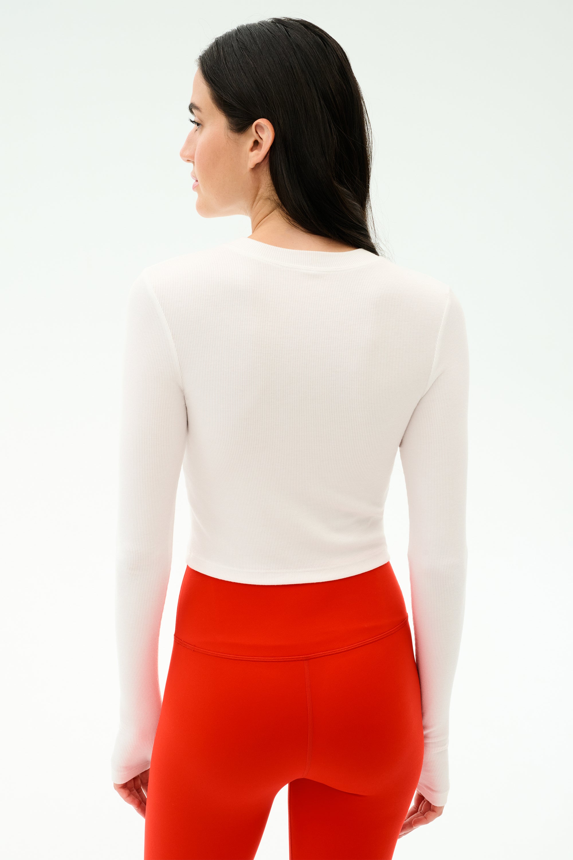 A woman with long dark hair models the SPLITS59 Louise Rib Long Sleeve Crop in white, paired with high-waisted red leggings, shown from the back against a plain white background.