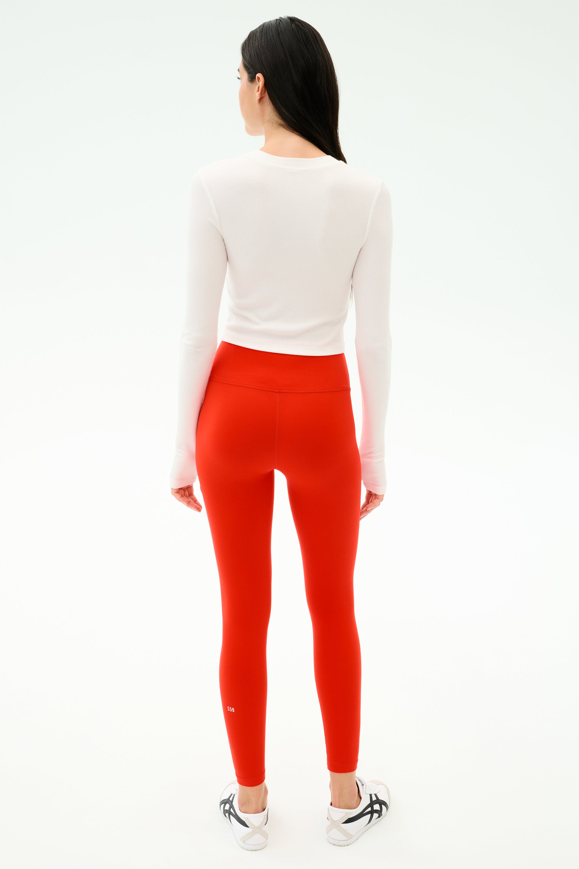 A person with long dark hair wears the SPLITS59 Louise Rib Long Sleeve Crop in White, paired with bright red leggings and white sneakers, standing with their back to the camera against a plain white background.