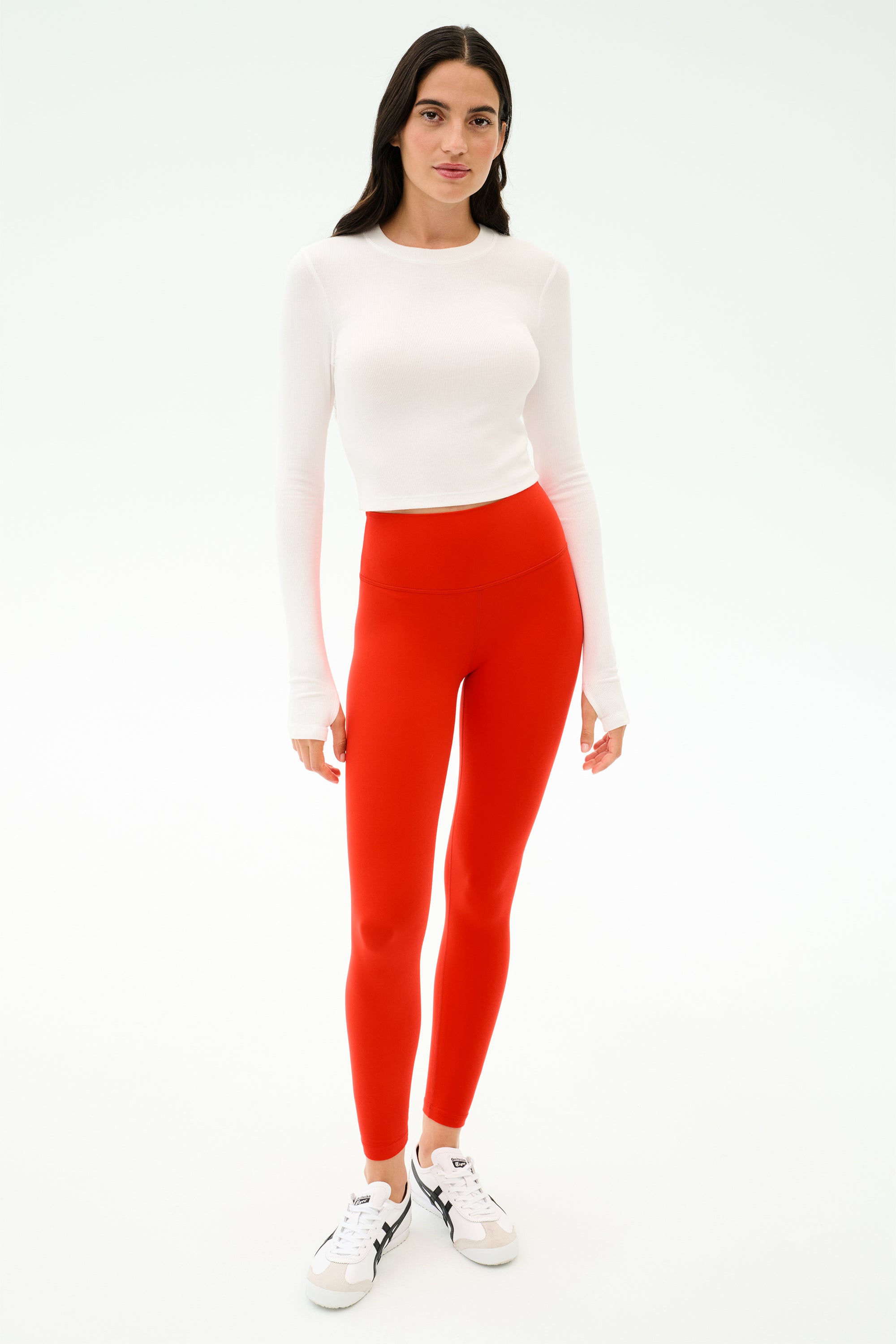 A woman stands against a white background wearing the SPLITS59 Louise Rib Long Sleeve Crop in white, paired with bright red leggings and white sneakers.