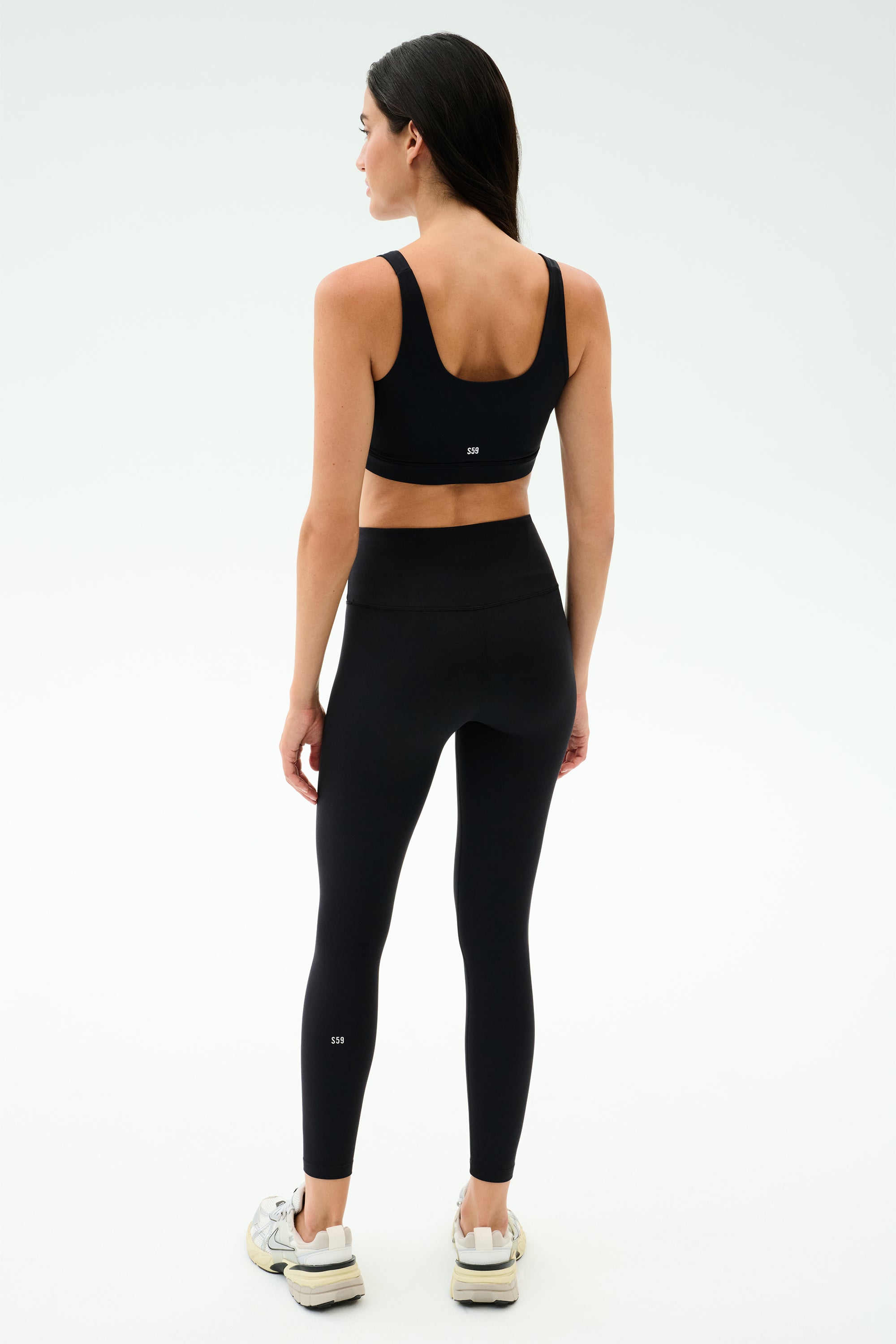 A woman faces away, wearing the SPLITS59 Sprint Rigor Bra in black, paired with black high-waisted leggings and white sneakers against a plain light background.