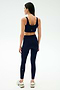 A woman stands with her back to the camera, wearing SPLITS59's Sprint High Waist Rigor 7/8 leggings in Indigo and a matching navy sports bra, paired with white sneakers against a plain light background.