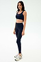 A woman stands neutrally in SPLITS59 Sprint High Waist Rigor 7/8 leggings in Indigo with a matching navy sports bra and white sneakers against a plain white background.