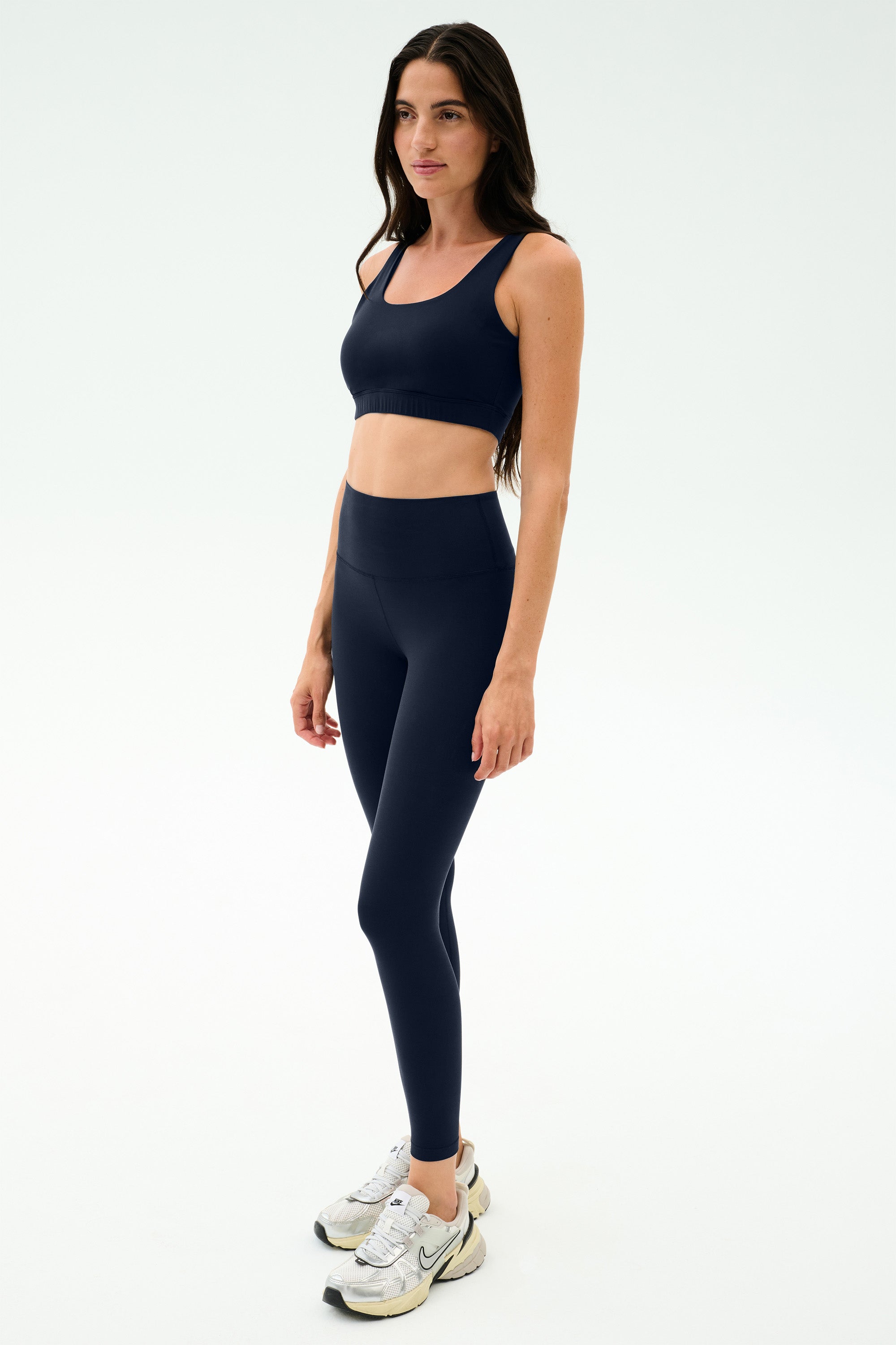 A woman stands neutrally in SPLITS59 Sprint High Waist Rigor 7/8 leggings in Indigo with a matching navy sports bra and white sneakers against a plain white background.