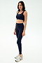 A woman stands neutrally in SPLITS59 Sprint High Waist Rigor 7/8 leggings in Indigo with a matching navy sports bra and white sneakers against a plain white background.