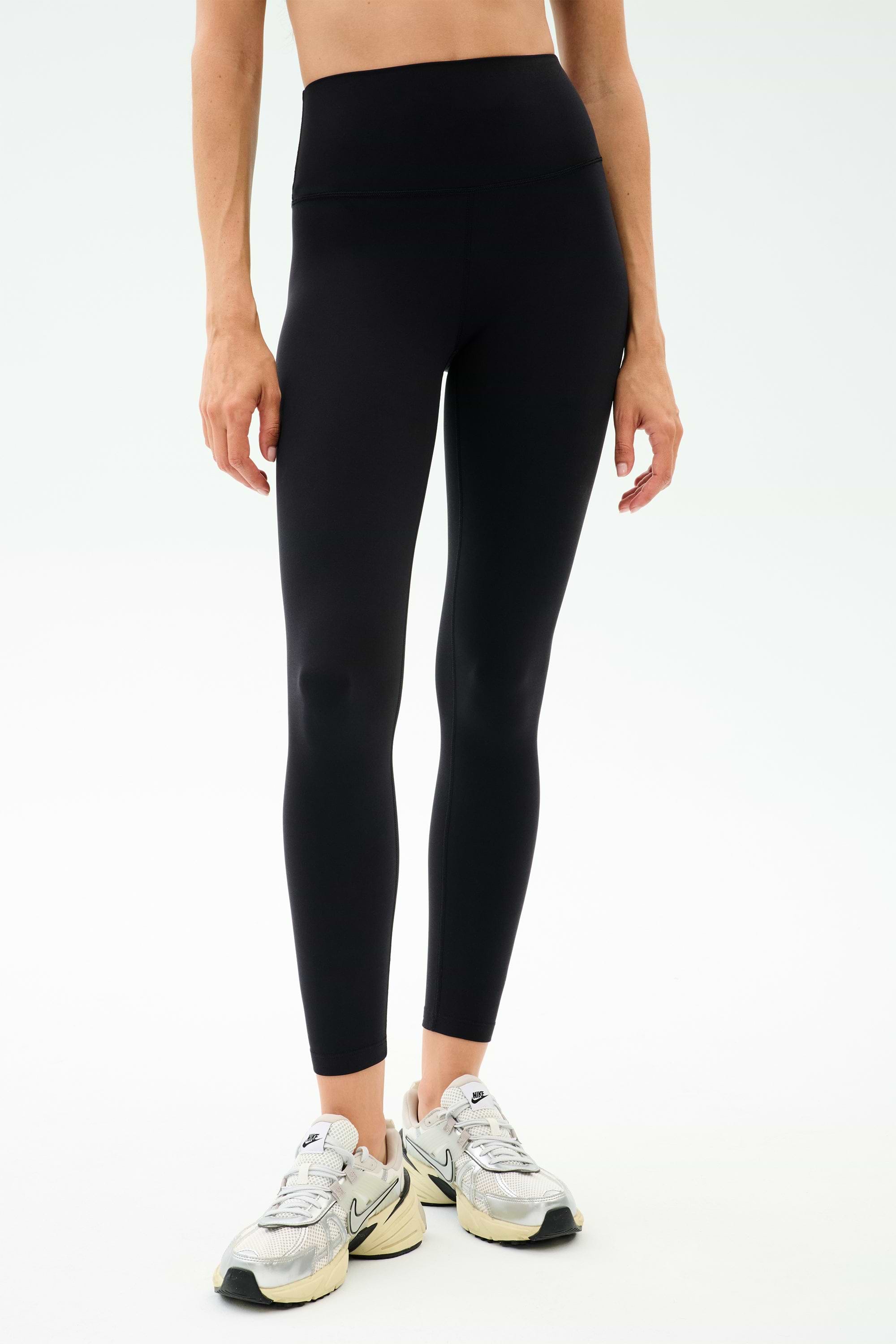 Model wearing SPLITS59 Sprint High Waist Rigor 7/8 leggings in black, paired with white athletic sneakers, shown from waist to ankles against a plain background.