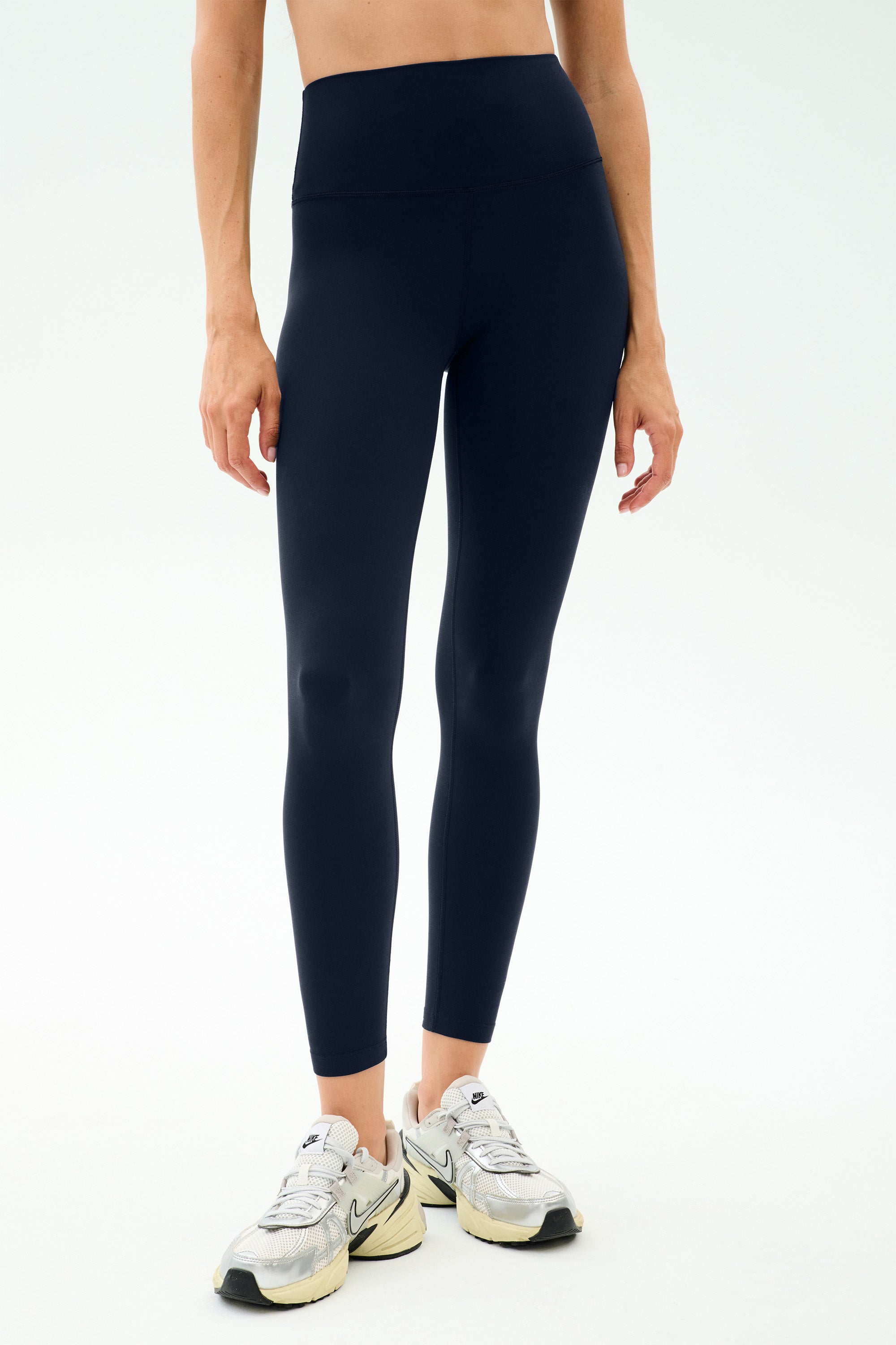 A person wearing SPLITS59 Sprint High Waist Rigor 7/8 leggings in Indigo with white athletic shoes stands against a plain light background.