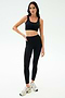 A woman stands against a plain background wearing SPLITS59's Sprint High Waist Rigor 7/8 leggings in black, paired with a sports bra and white sneakers.