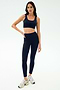 A woman stands against a plain background wearing SPLITS59 Sprint High Waist Rigor 7/8 leggings in Indigo, paired with a matching sports bra and white running shoes.