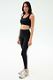 A woman stands against a plain white background wearing the SPLITS59 Sprint Rigor Bra in black, paired with black leggings and white sneakers, ready for her gym workout.