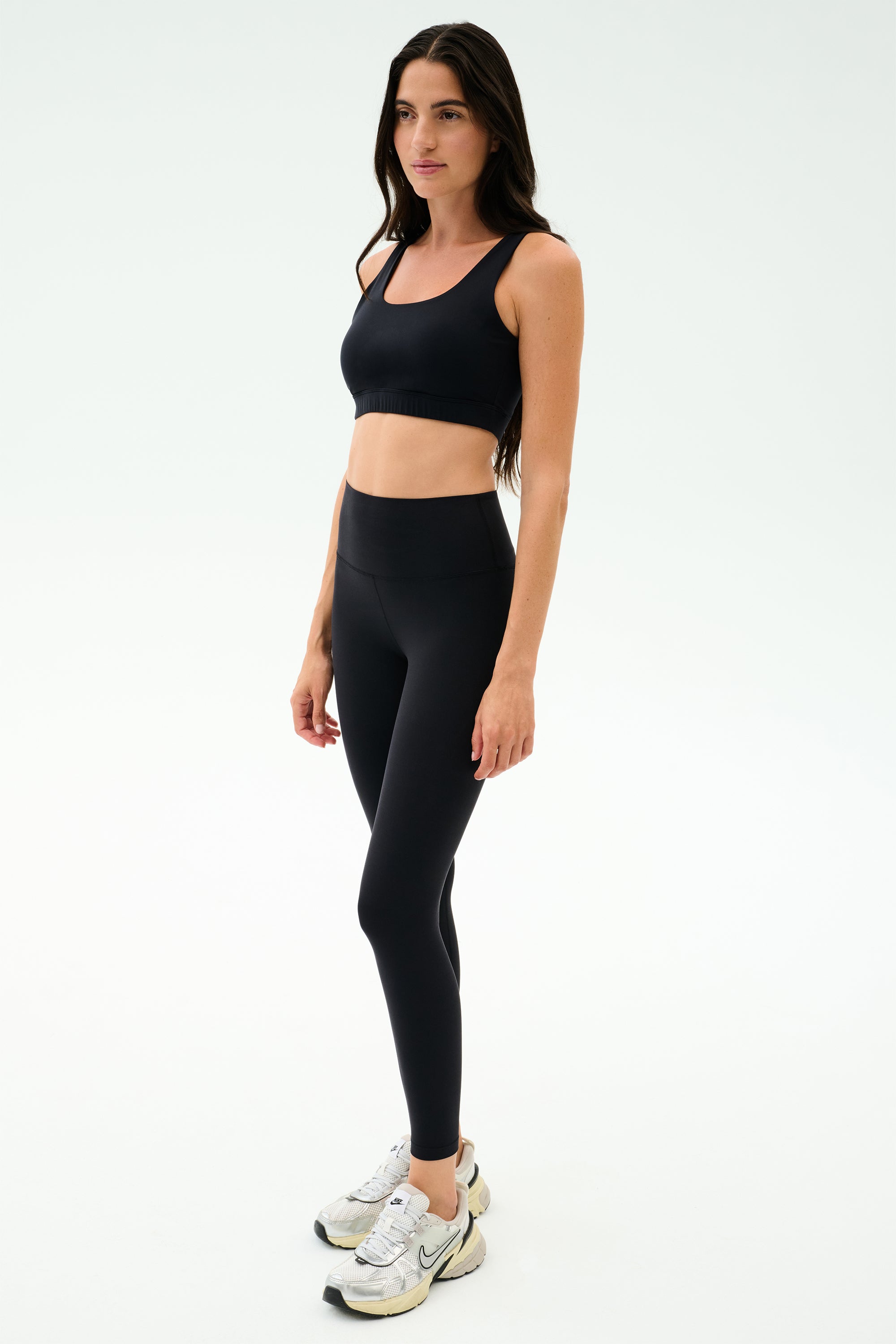 A woman stands against a plain white background wearing the SPLITS59 Sprint Rigor Bra in black, paired with black leggings and white sneakers, ready for her gym workout.