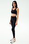 A woman stands against a plain white background wearing the SPLITS59 Sprint Rigor Bra in black, paired with black leggings and white sneakers, ready for her gym workout.