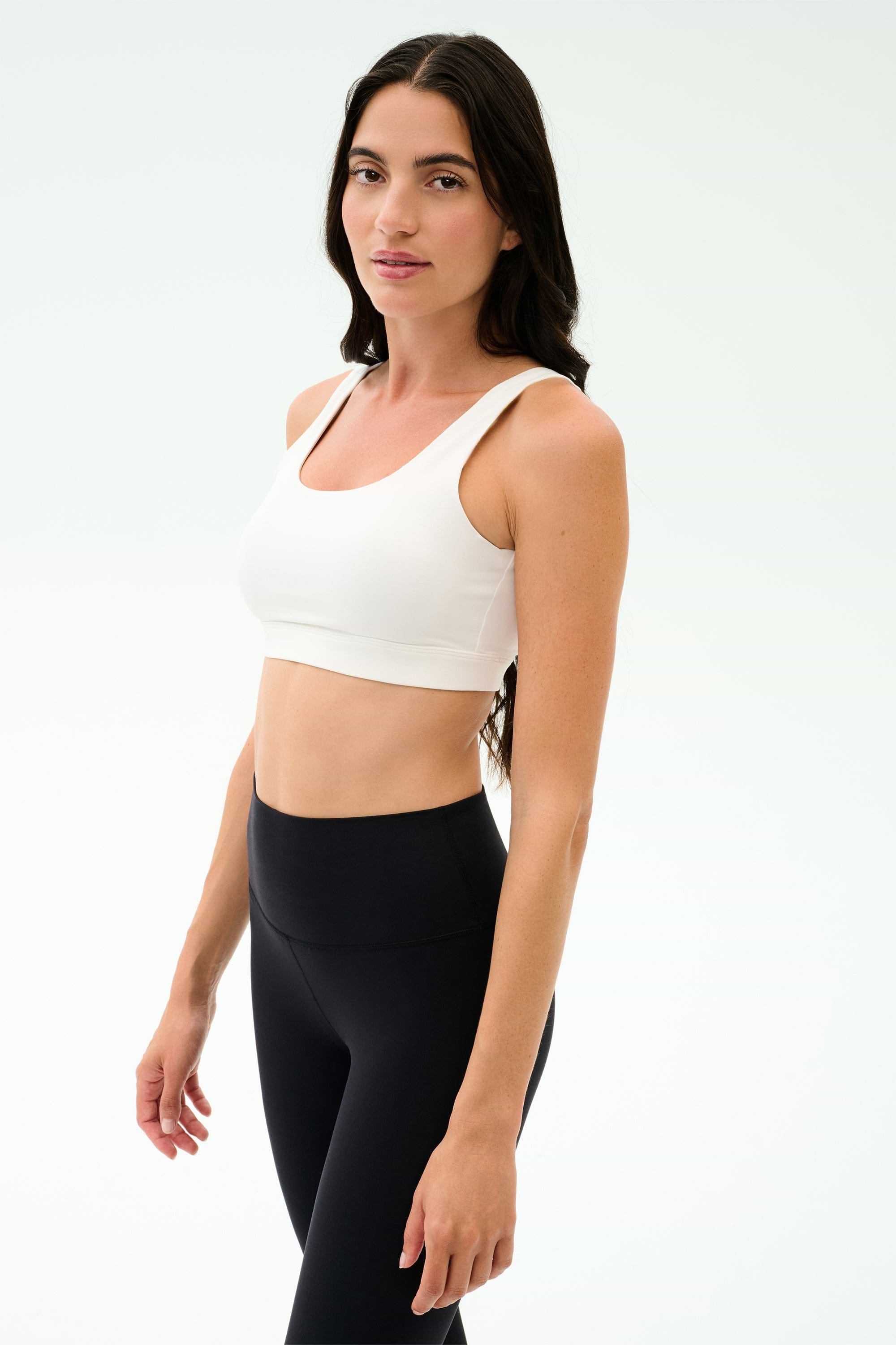 Woman with long dark hair wears the SPLITS59 Sprint Rigor Bra in white and black high-waisted leggings, standing in a well-lit studio with a neutral background—perfect for gym sessions and versatile multi-sport performance.