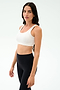 Woman with long dark hair wears the SPLITS59 Sprint Rigor Bra in white and black high-waisted leggings, standing in a well-lit studio with a neutral background—perfect for gym sessions and versatile multi-sport performance.