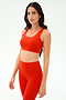 Woman stands against a plain background, wearing SPLITS59’s Sprint Rigor Bra in Lotus Red with matching leggings, looking at the camera—ideal for multisport performance and gym workouts.