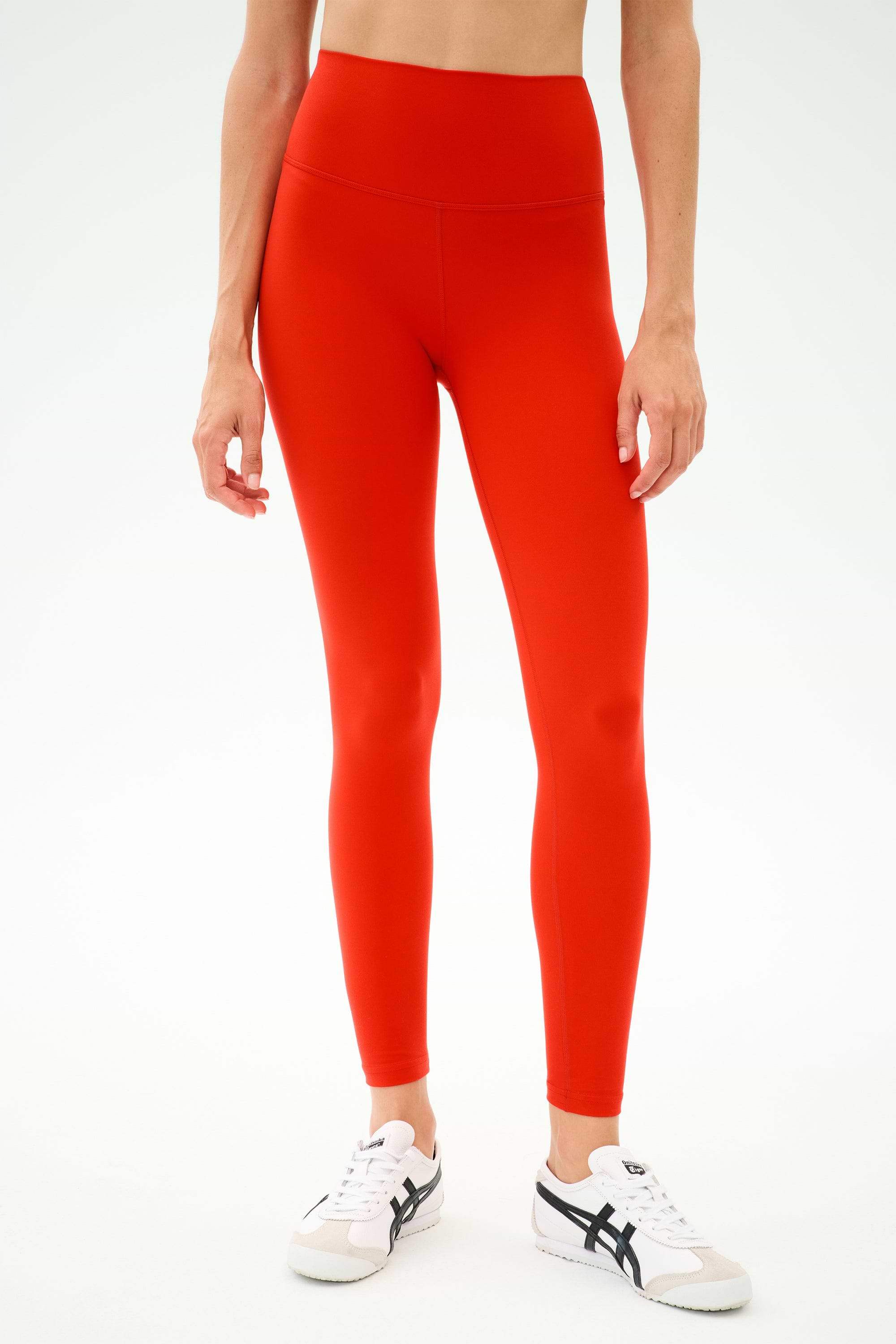 A person wearing SPLITS59 Sprint High Waist Rigor 7/8 leggings in Lotus Red with white sneakers stands against a plain light background.