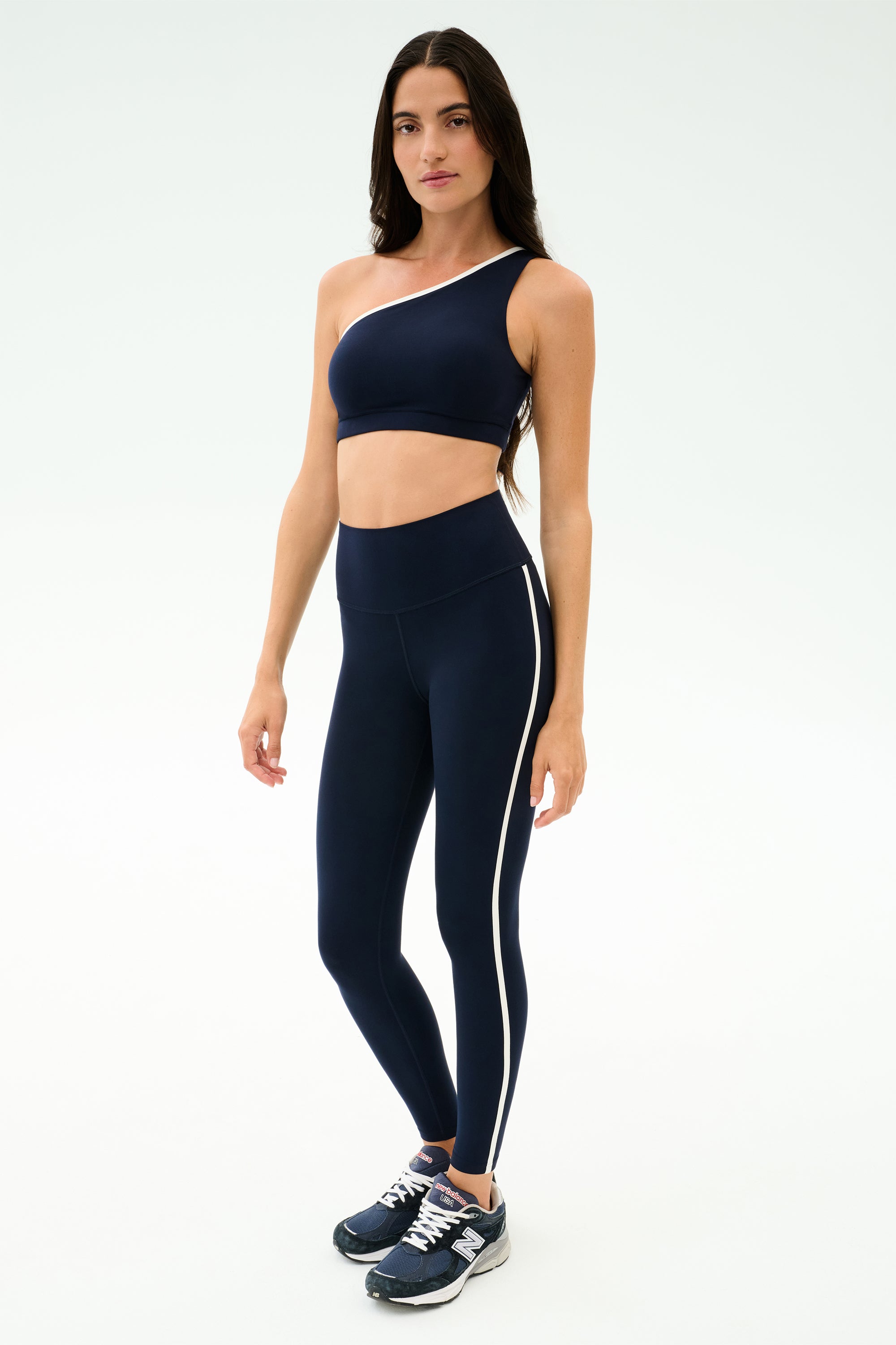 A woman wears the SPLITS59 Amber Airweight High Waist 7/8 leggings in Indigo/White—featuring a one-shoulder sports bra and dark athletic shoes—against a plain light background, ideal for hot yoga.