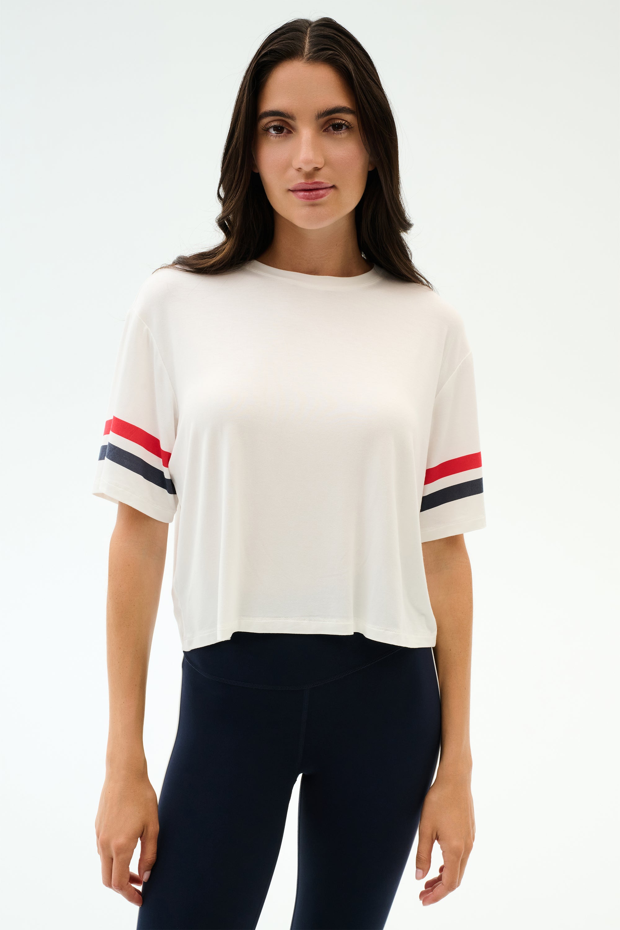 A woman faces forward in the SPLITS59 Ava Jersey Tee - White/Indigo, featuring a relaxed fit and sporty indigo and red sleeve stripes, paired with black leggings, set against a plain light background.