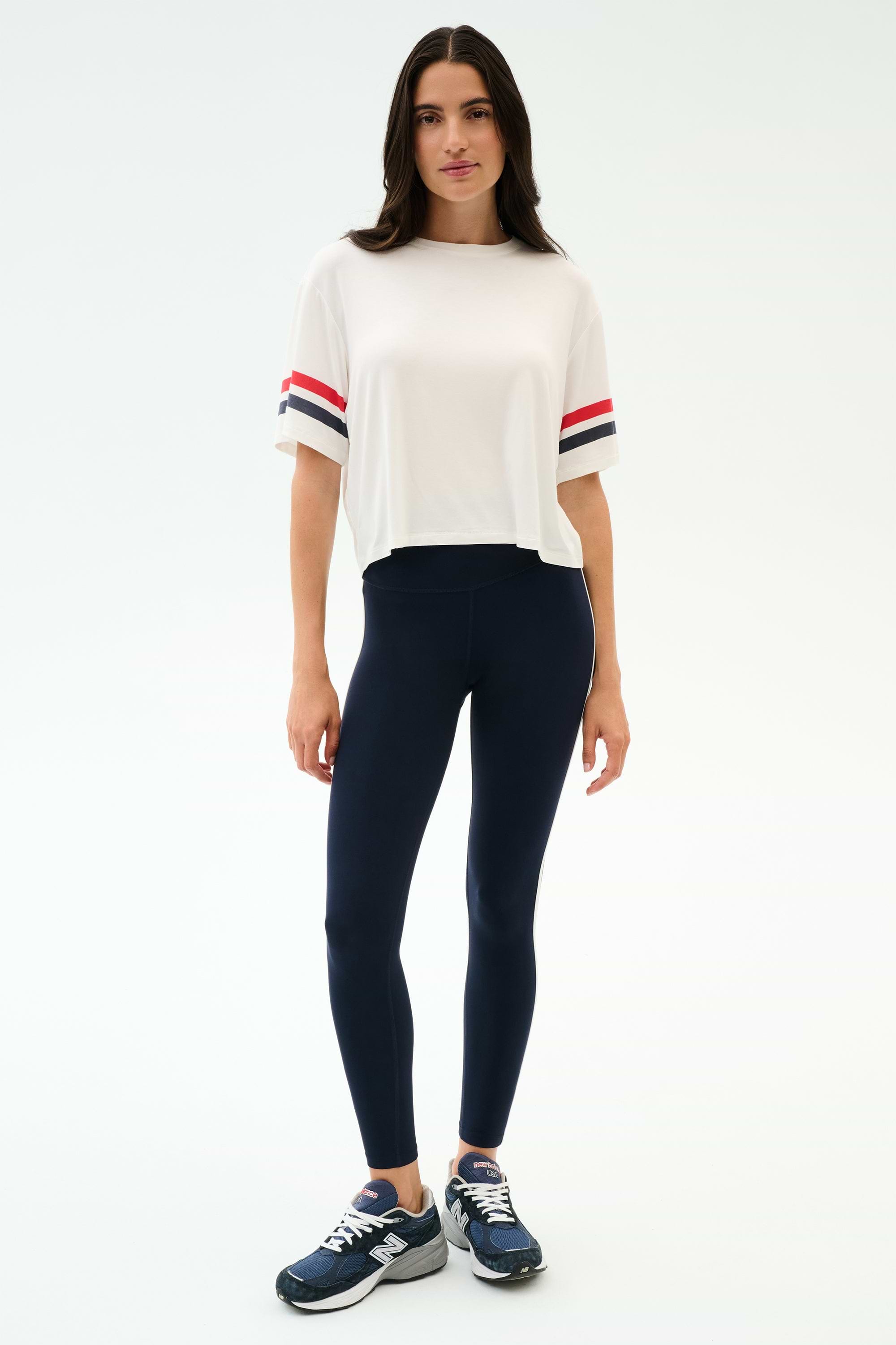 A woman wears the SPLITS59 Ava Jersey Tee in White/Indigo, featuring a relaxed fit and sporty sleeve stripes, paired with blue leggings—perfect for gym workouts.