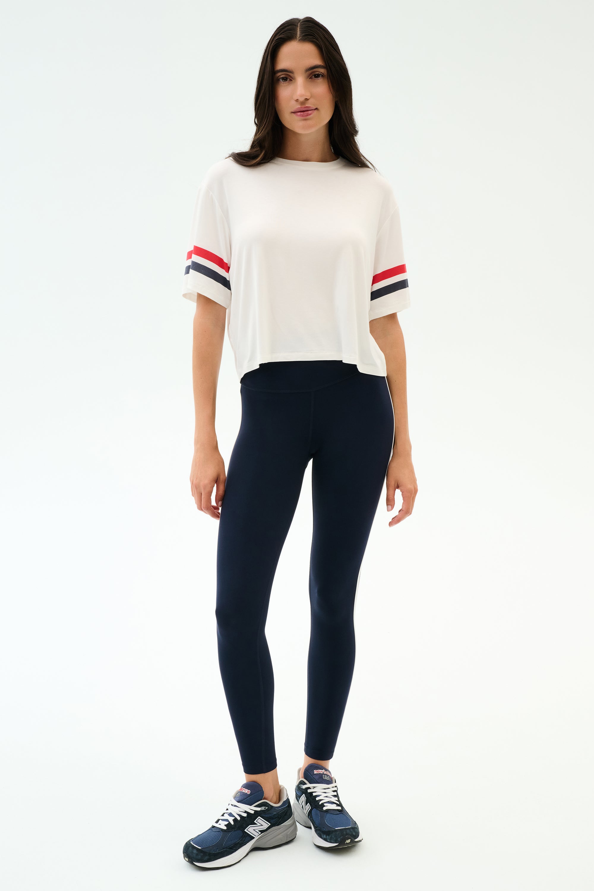 A woman wears the SPLITS59 Ava Jersey Tee in White/Indigo, featuring a relaxed fit and sporty sleeve stripes, paired with blue leggings—perfect for gym workouts.