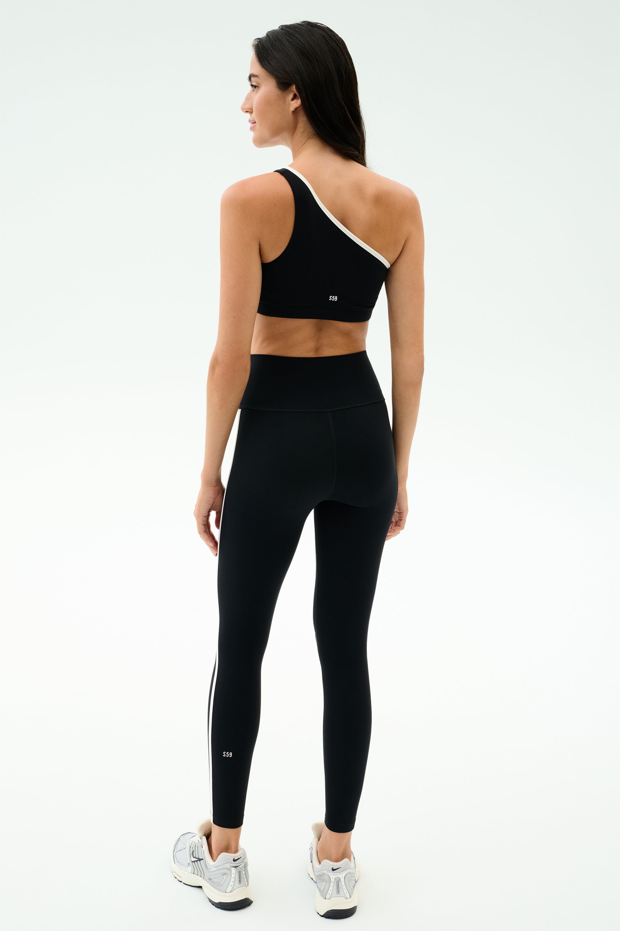 A woman stands facing away on a light background, wearing the SPLITS59 Josie Airweight One Shoulder Bra in black/white with matching black leggings featuring white side stripes, paired with white athletic shoes.
