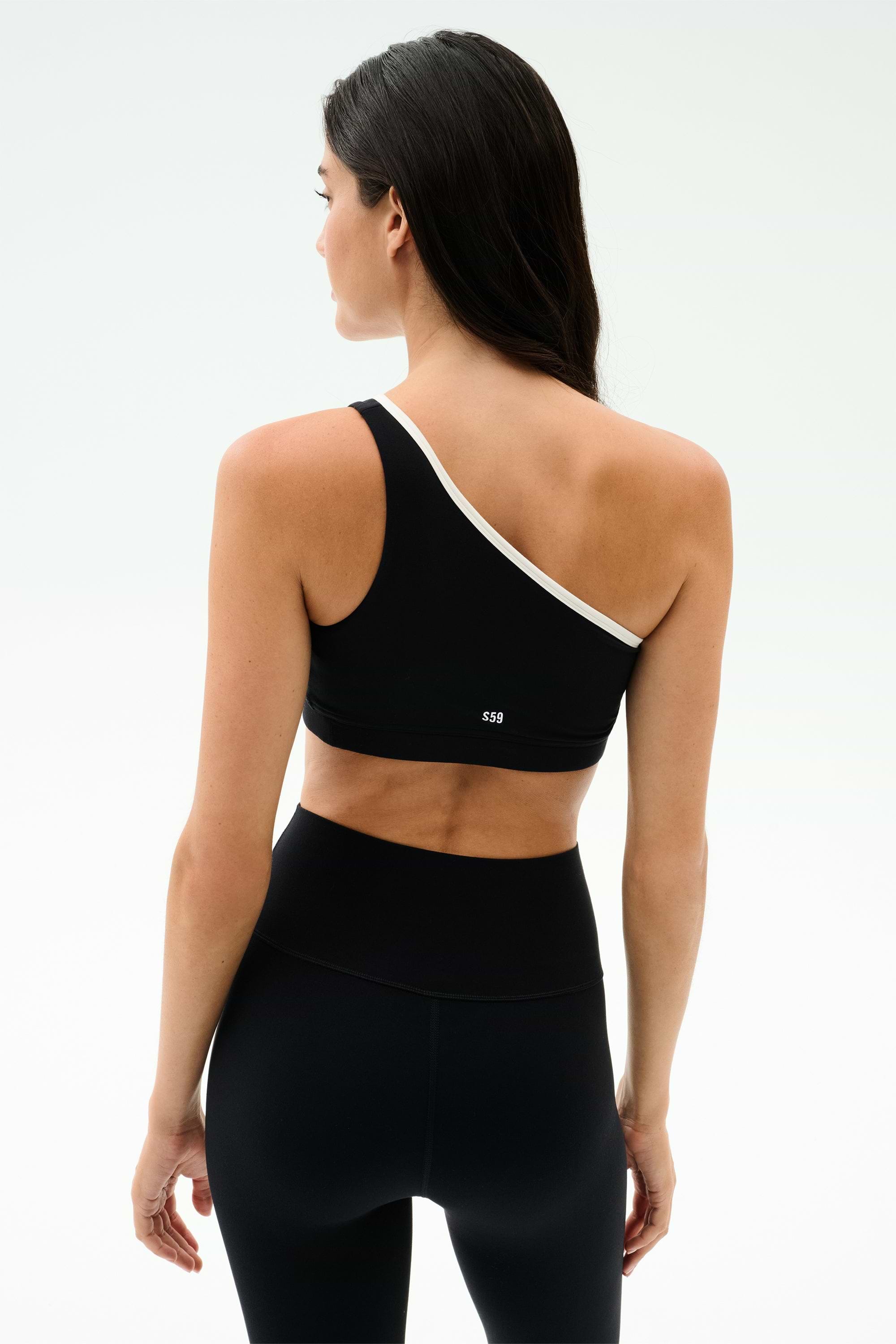 A woman faces away, wearing the SPLITS59 Josie Airweight One Shoulder Bra in Black/White with matching high-waisted leggings, set against a plain light background.