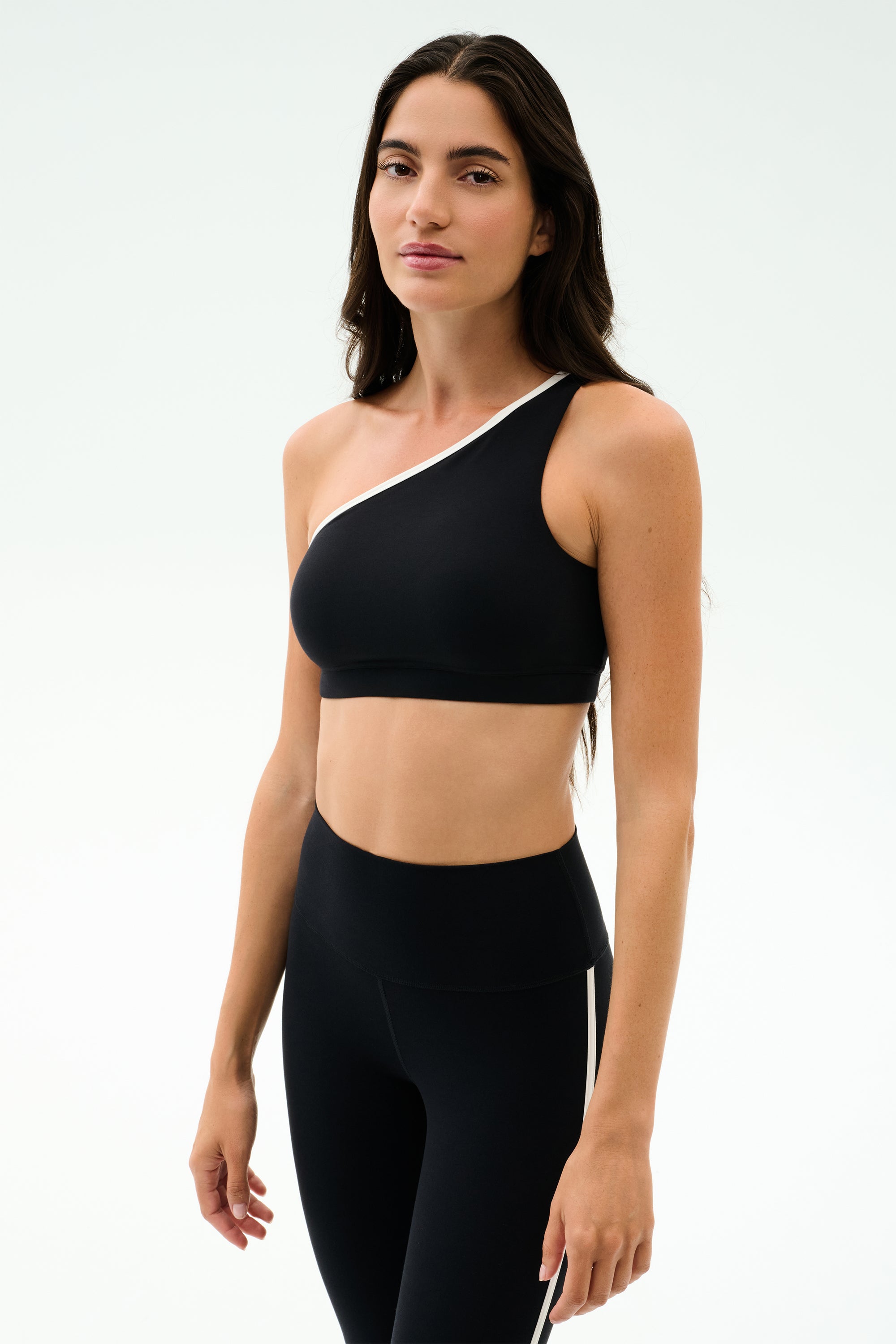 A woman models the SPLITS59 Josie Airweight One Shoulder Bra in Black/White with matching leggings, ideal for hot yoga, against a simple light backdrop.