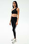 A woman wears the SPLITS59 Josie Airweight One Shoulder Bra in Black/White with black leggings featuring a white side stripe and white athletic shoes, standing against a plain light background—ideal for hot yoga sessions.