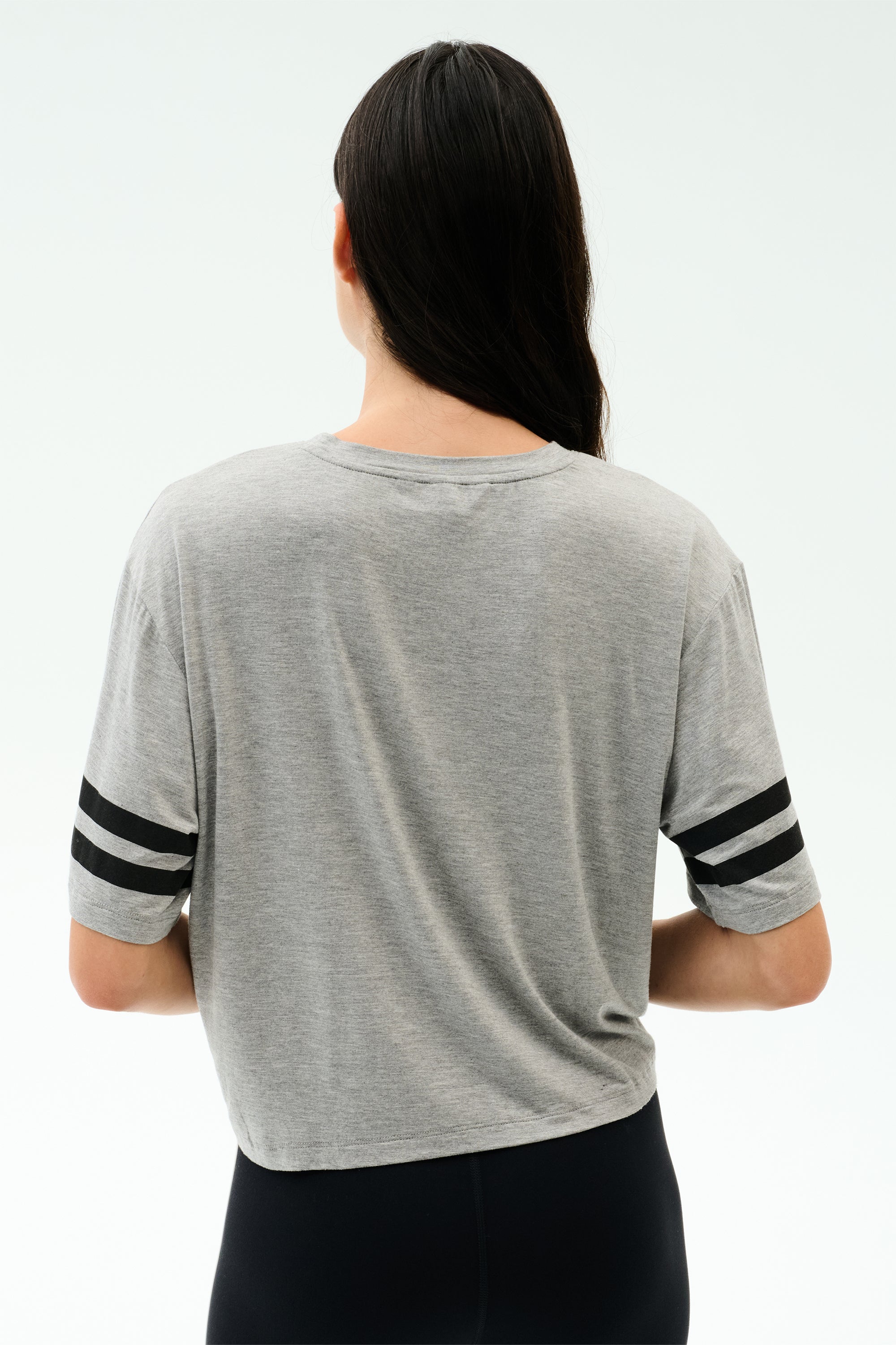 A person with long dark hair, seen from the back, wears the SPLITS59 Ava Jersey Tee in Heather Grey/Black and black pants against a plain light backdrop—perfect for gym sessions or CrossFit.