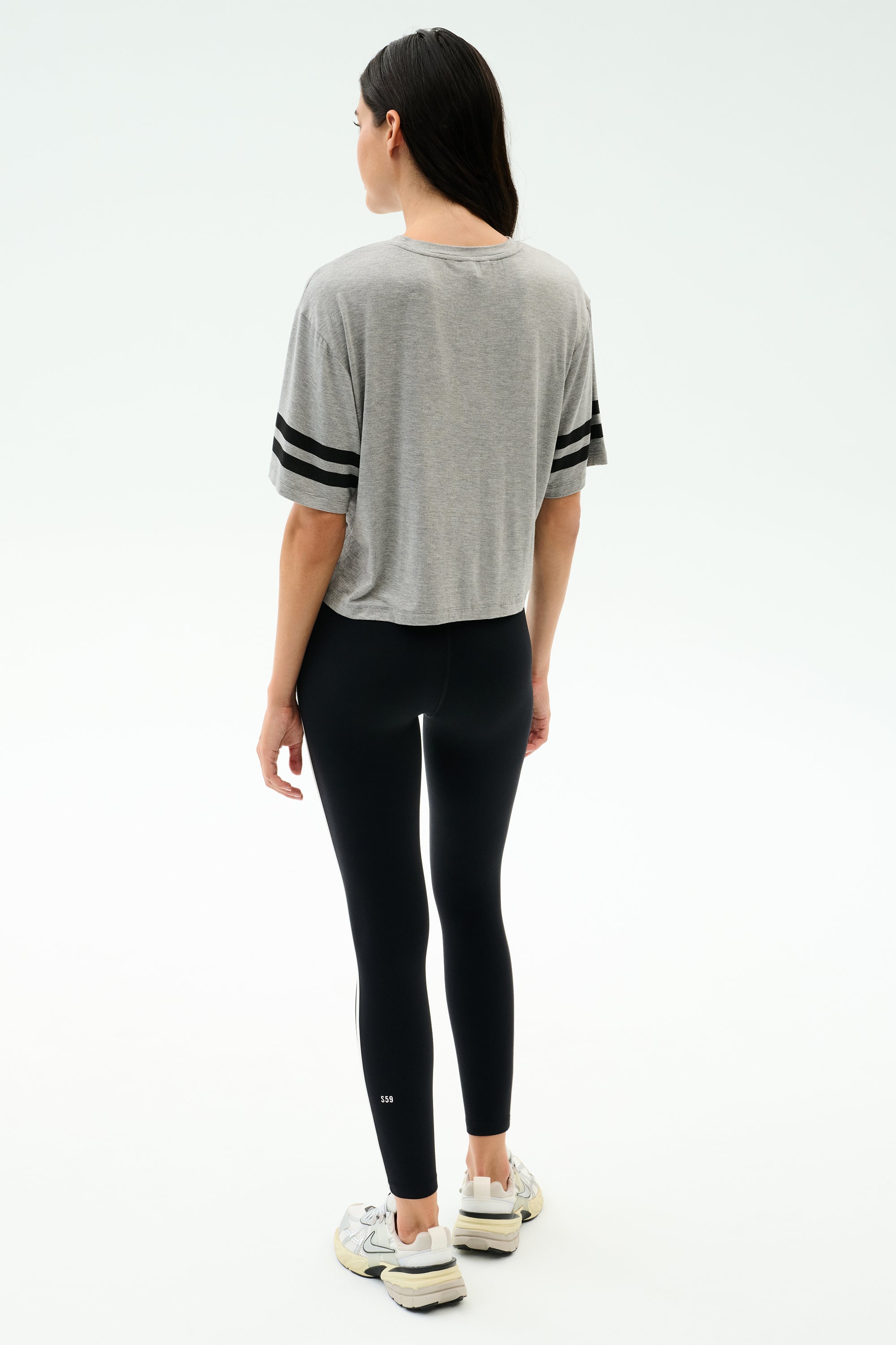 A woman faces away, wearing the SPLITS59 Ava Jersey Tee in Heather Grey/Black, paired with black leggings and white sneakers, set against a plain light background.