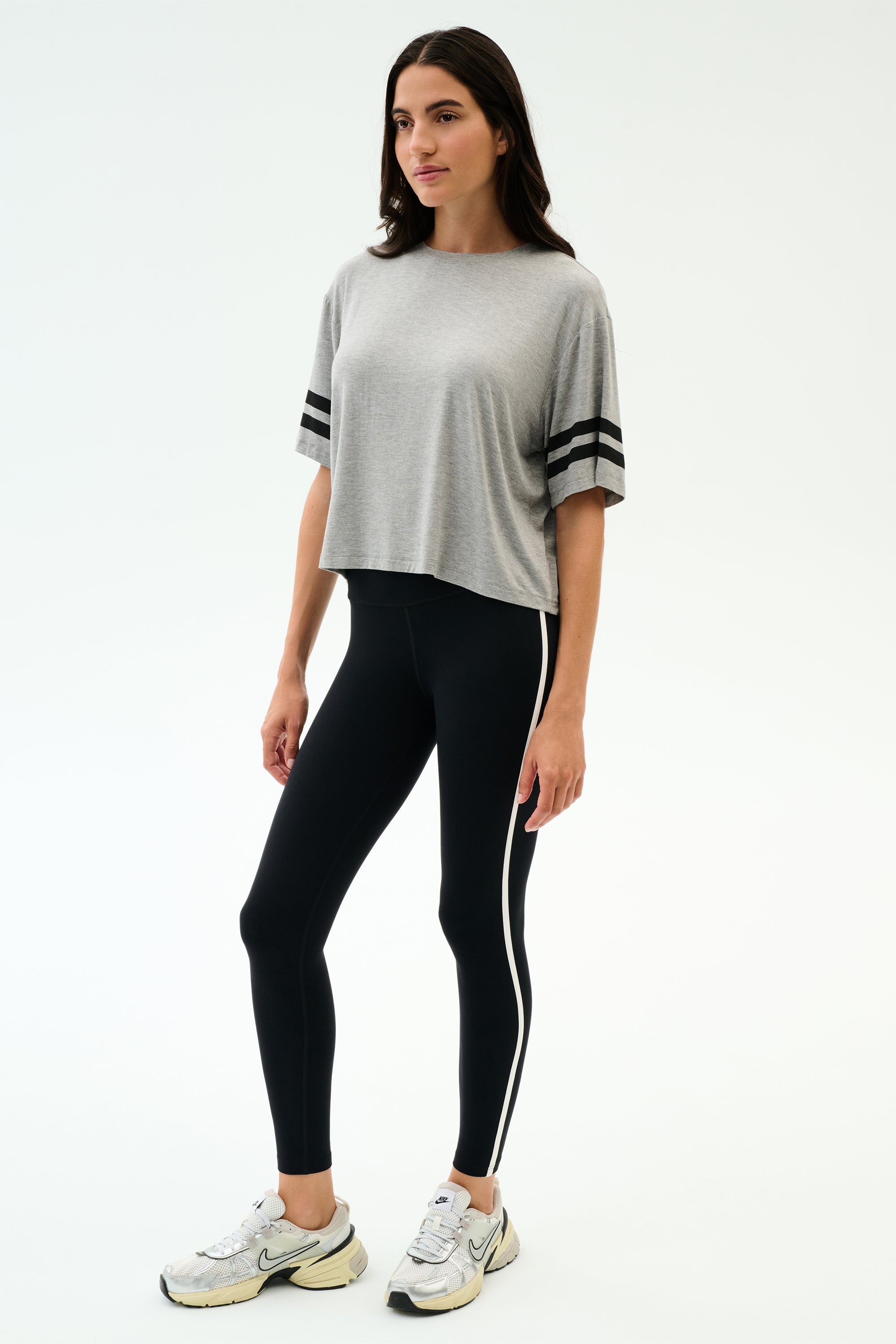 A woman stands against a plain background wearing the SPLITS59 Ava Jersey Tee in Heather Grey/Black, paired with black leggings with a white side stripe and white sneakers, ready for her gym workout.